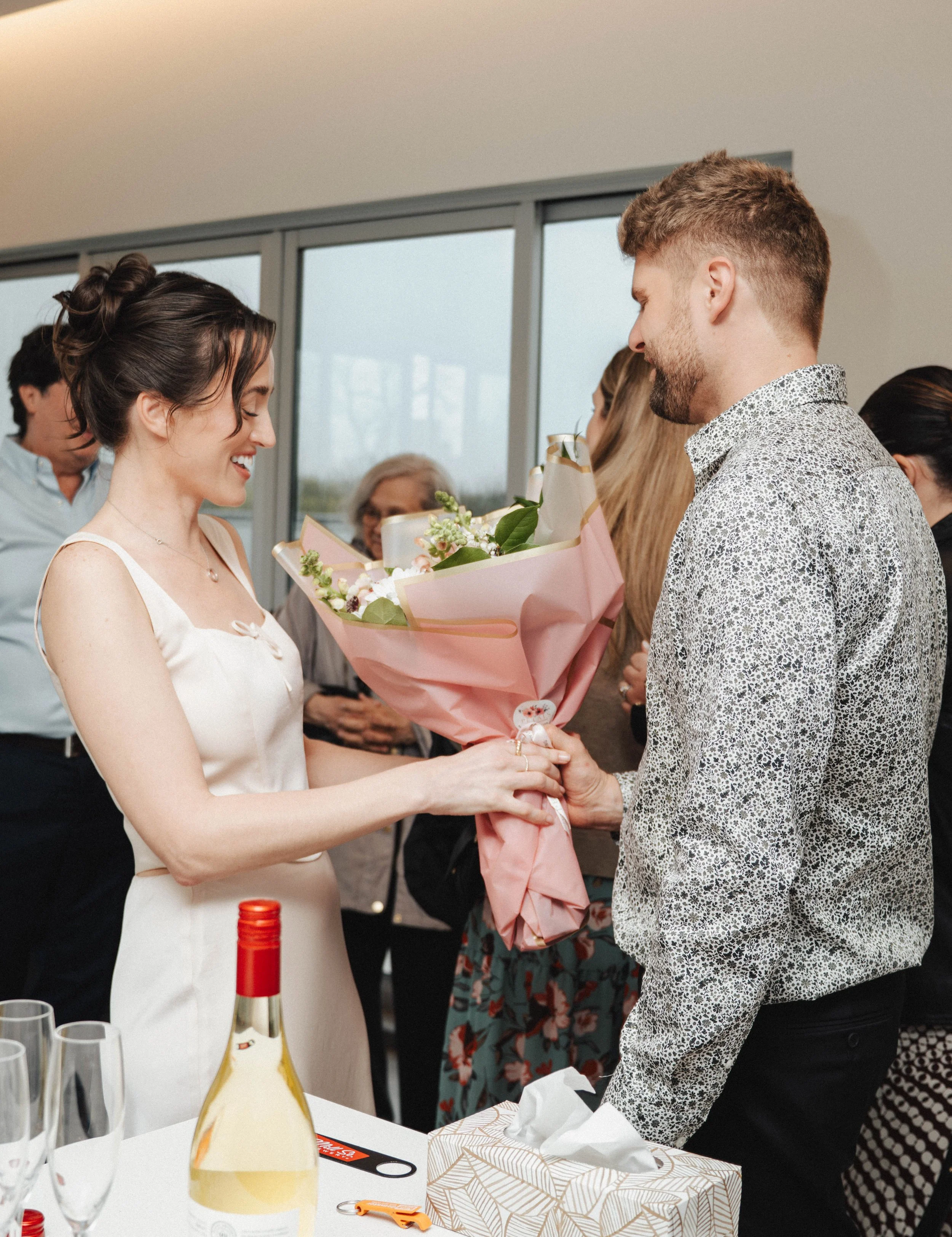 A woman in a white dress is receiving a bouquet of flowers from a man at a celebratory event. The scene appears to be joyful, with people in the background and items like a bottle of wine, glasses, a tissue box, and a gift on the table.