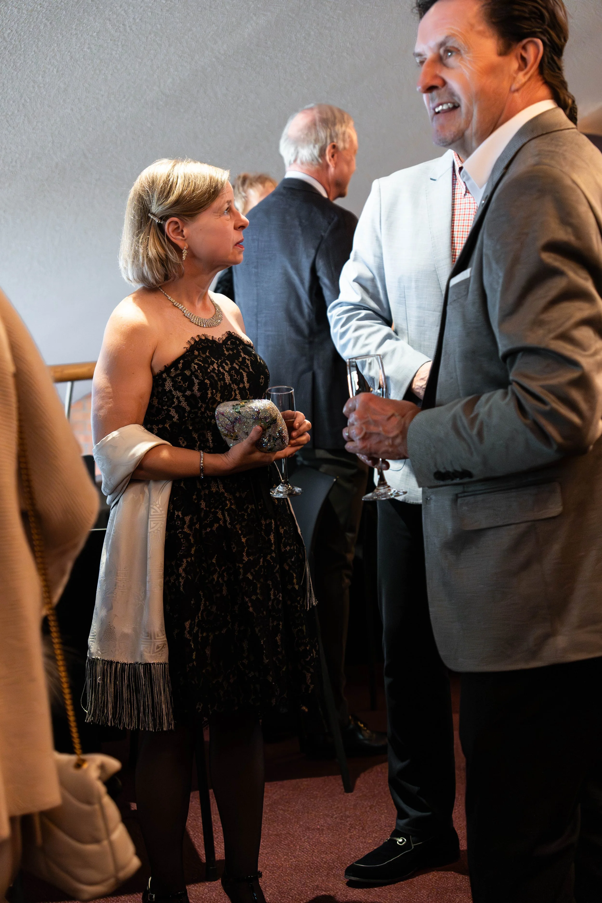 A woman in a black lace dress holding a decorated clutch and a glass of champagne, talking to a man in a light gray suit holding a glass of champagne, at a social event.