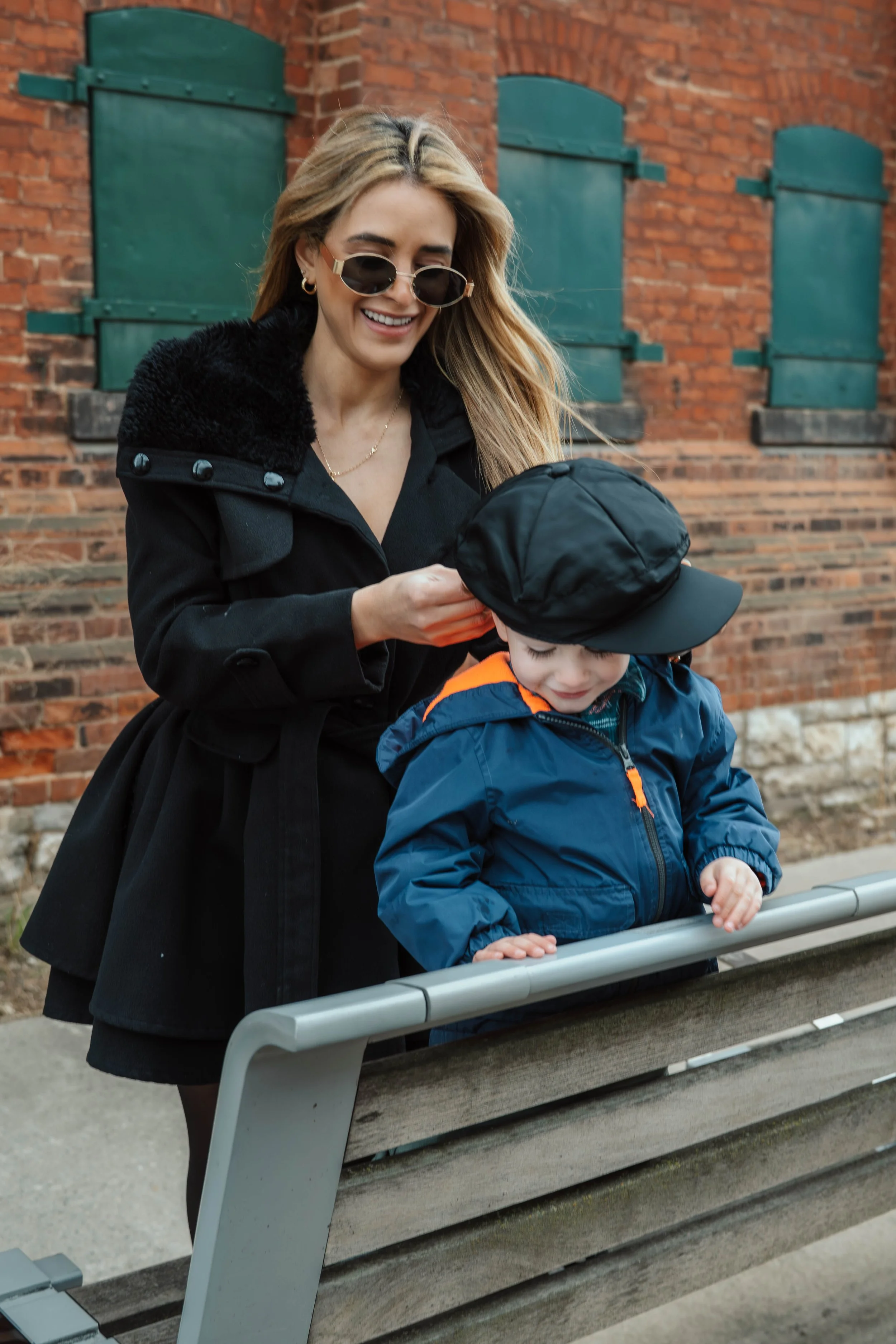 A woman with blonde hair wearing sunglasses and a black coat smiling while looking at a young boy in a blue jacket and black cap, standing at a park with a brick wall and green shutters in the background.