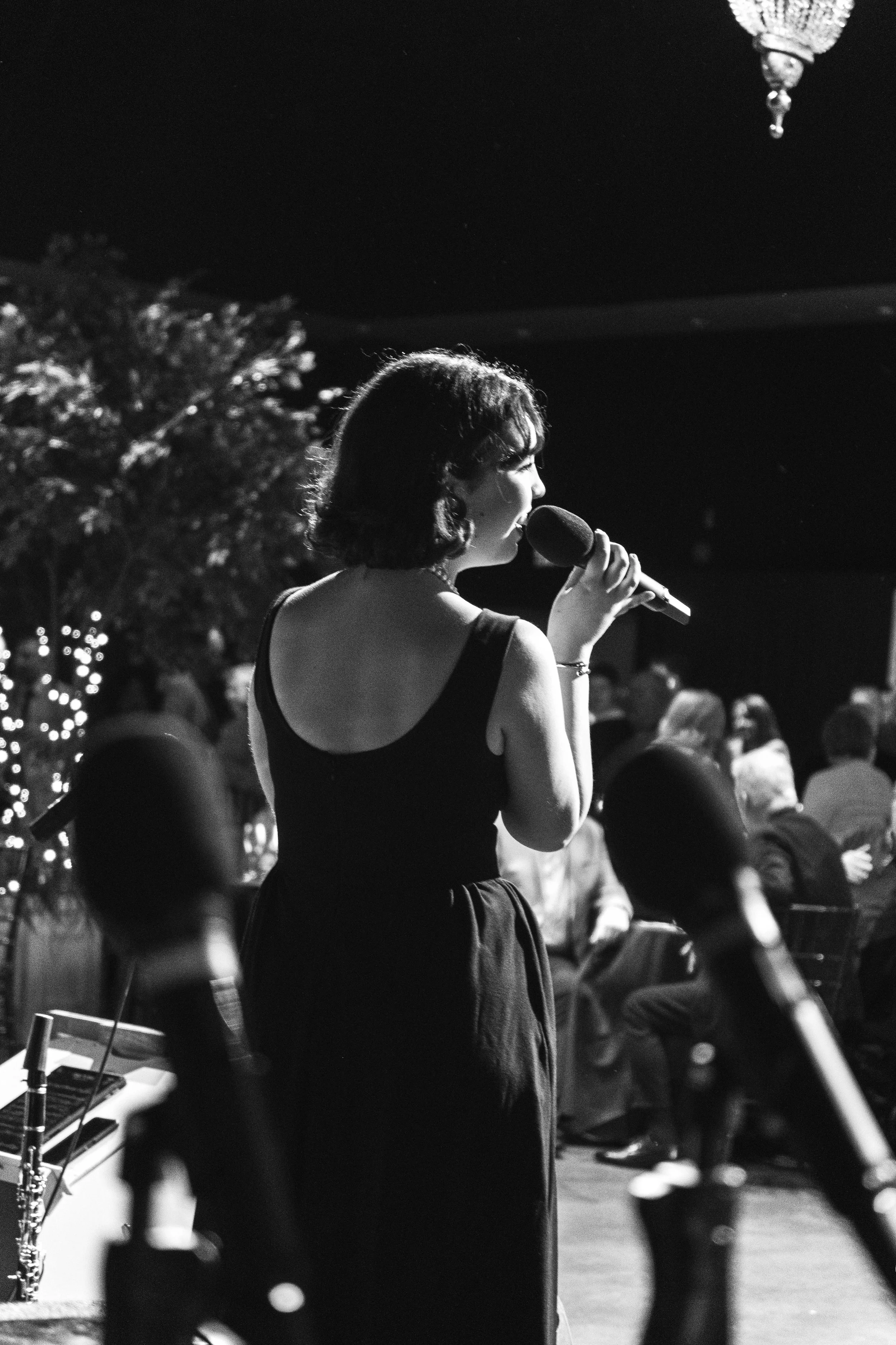 A woman singing into a microphone at a performance event, with an audience in the background, indoor setting, black and white photo.