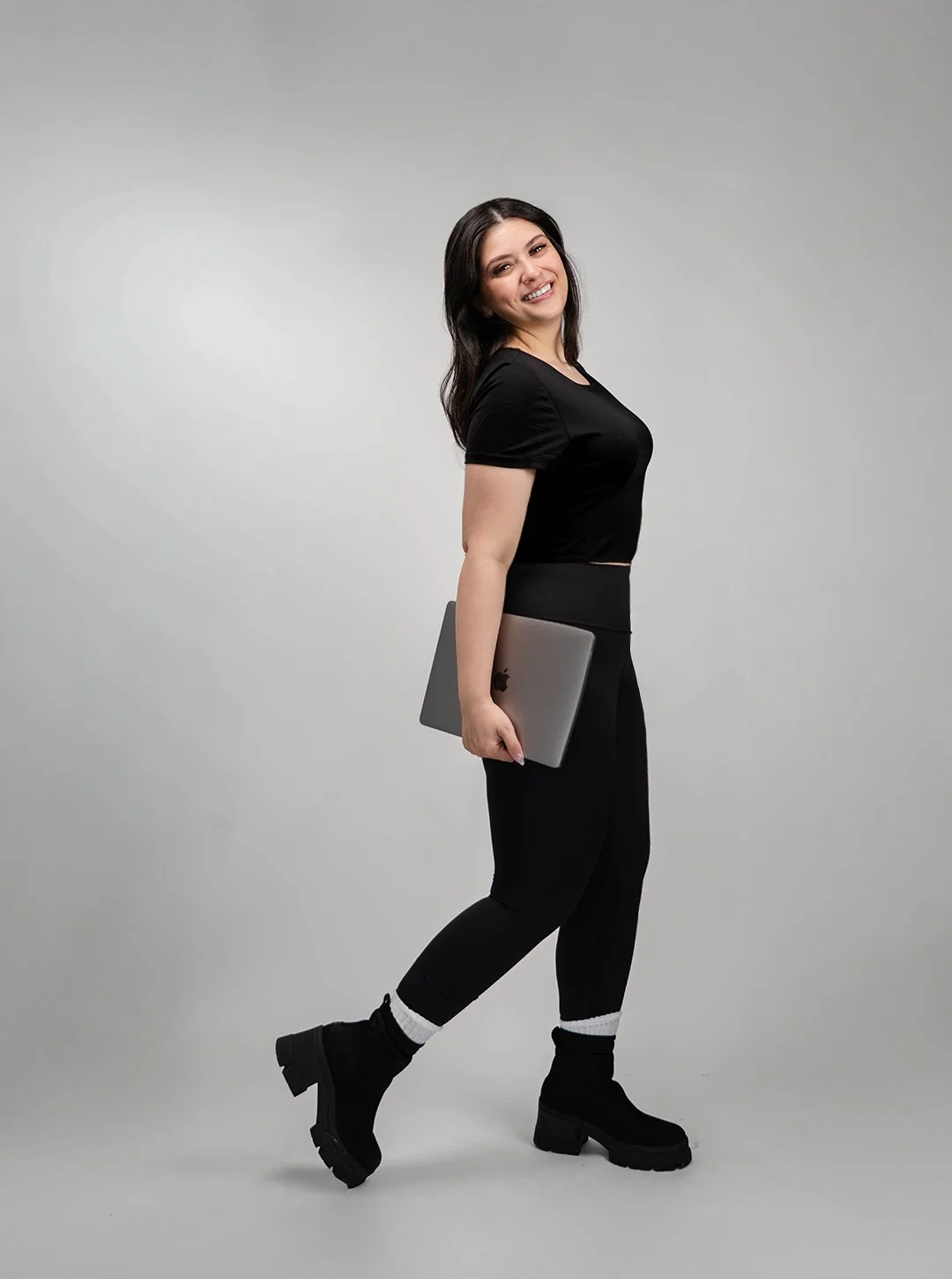 A young woman with dark hair, smiling, holding a laptop, wearing a black t-shirt, black pants, and black boots, standing against a plain light gray background.
