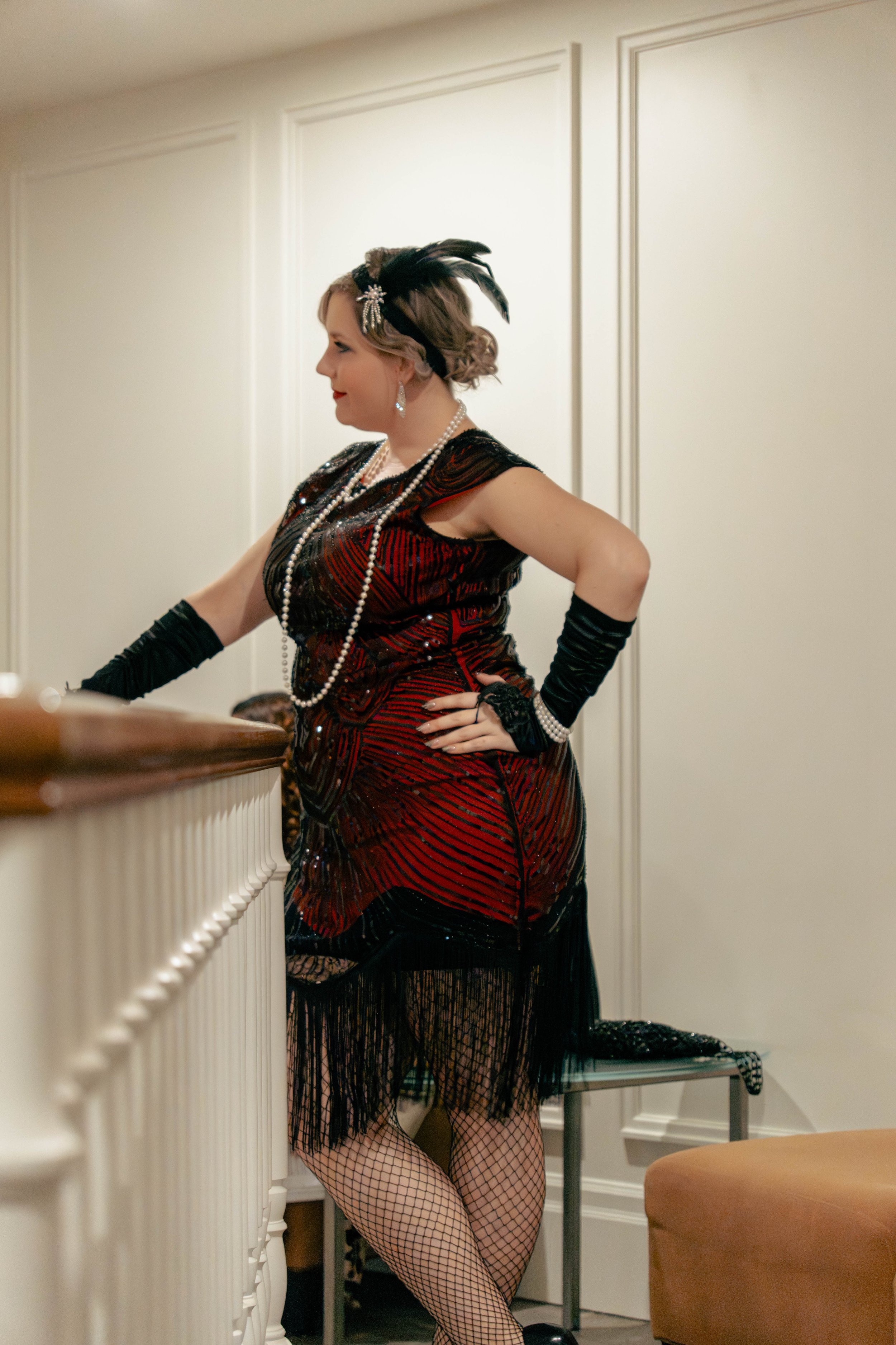 A woman dressed in vintage 1920s style attire, including a red and black flapper dress with fringe, fishnet stockings, black gloves, and a feathered headpiece, standing indoors with her hand on her hip.
