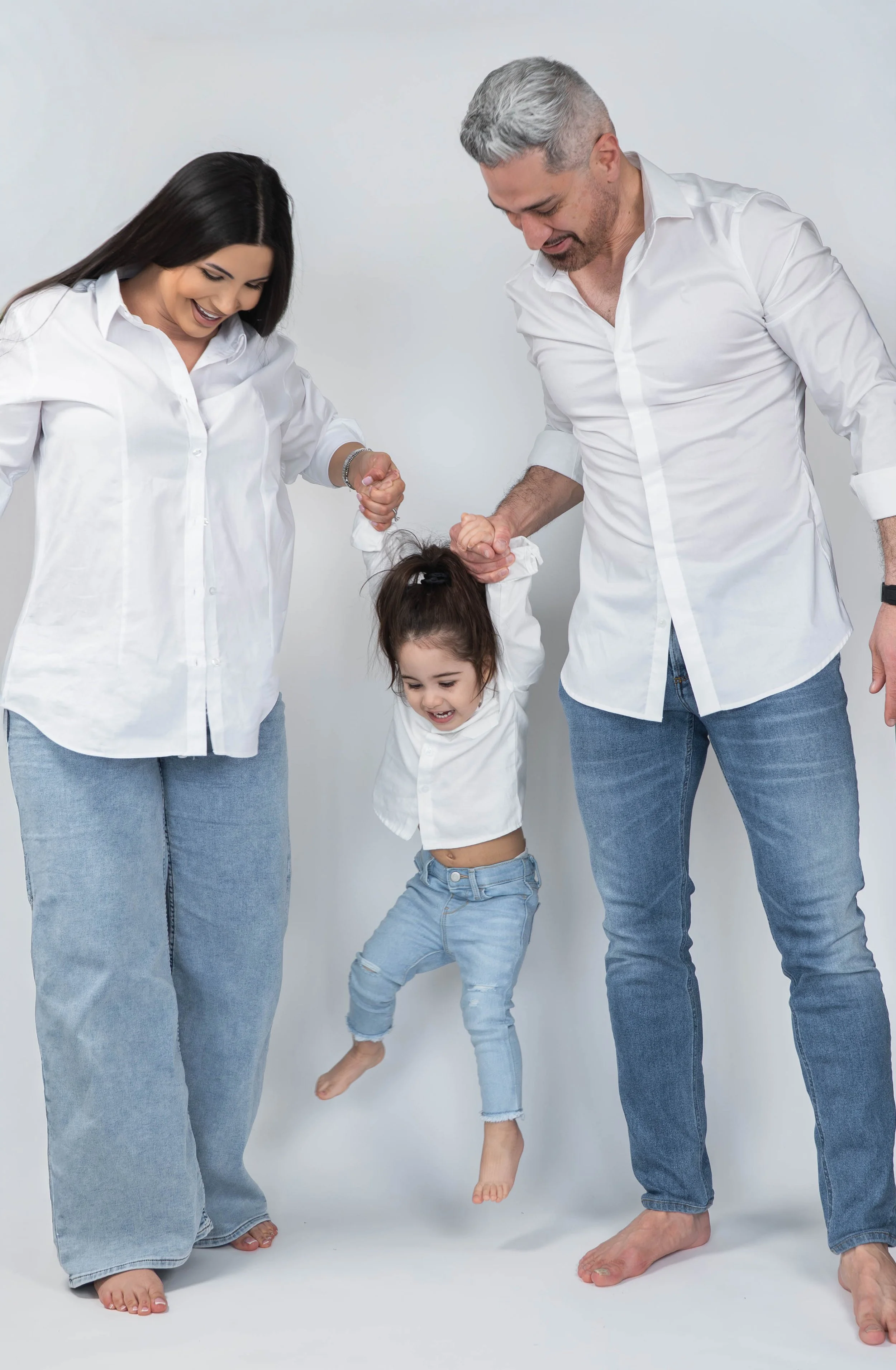 A family of four with light skin, including a woman, a man, and two children, standing together against a white background. The woman and man are holding hands with the children, and the little girl is mid-air, jumping or playing together with them. 