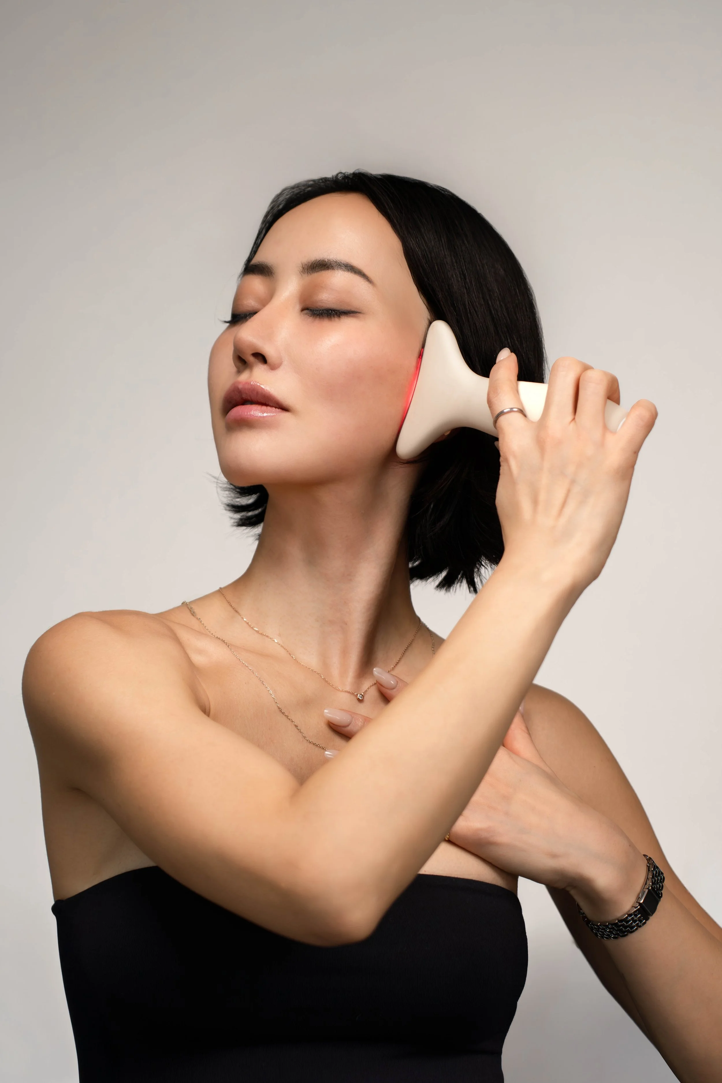 A woman with black hair is using a handheld device on her face with her eyes closed.