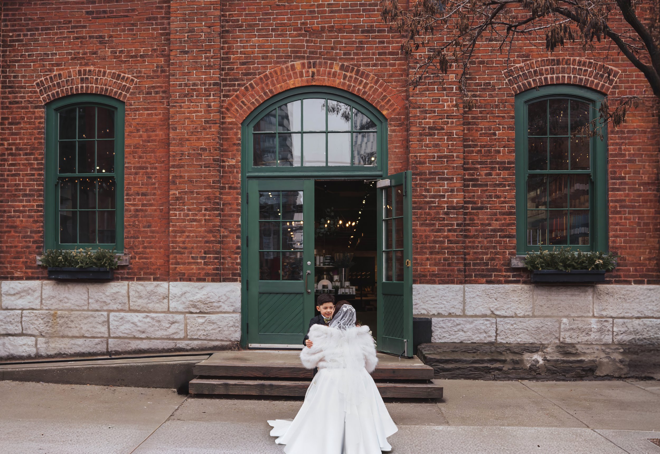 A young boy and girl dressed in formal attire, possibly for a wedding, standing outside a brick building with large green windows and open green door.