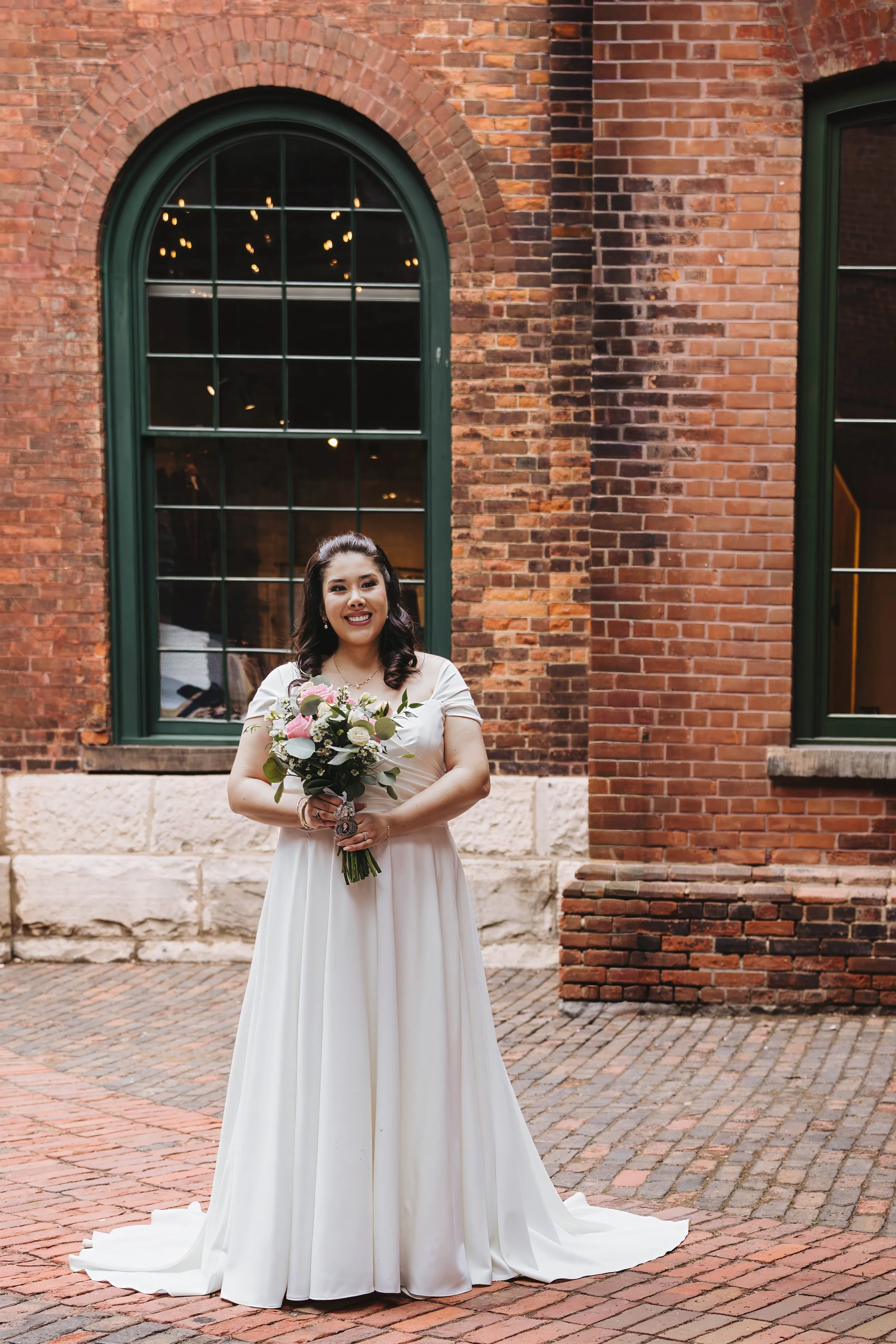A woman in a white wedding dress holding a bouquet of flowers, standing in front of a brick building with large arched green windows.