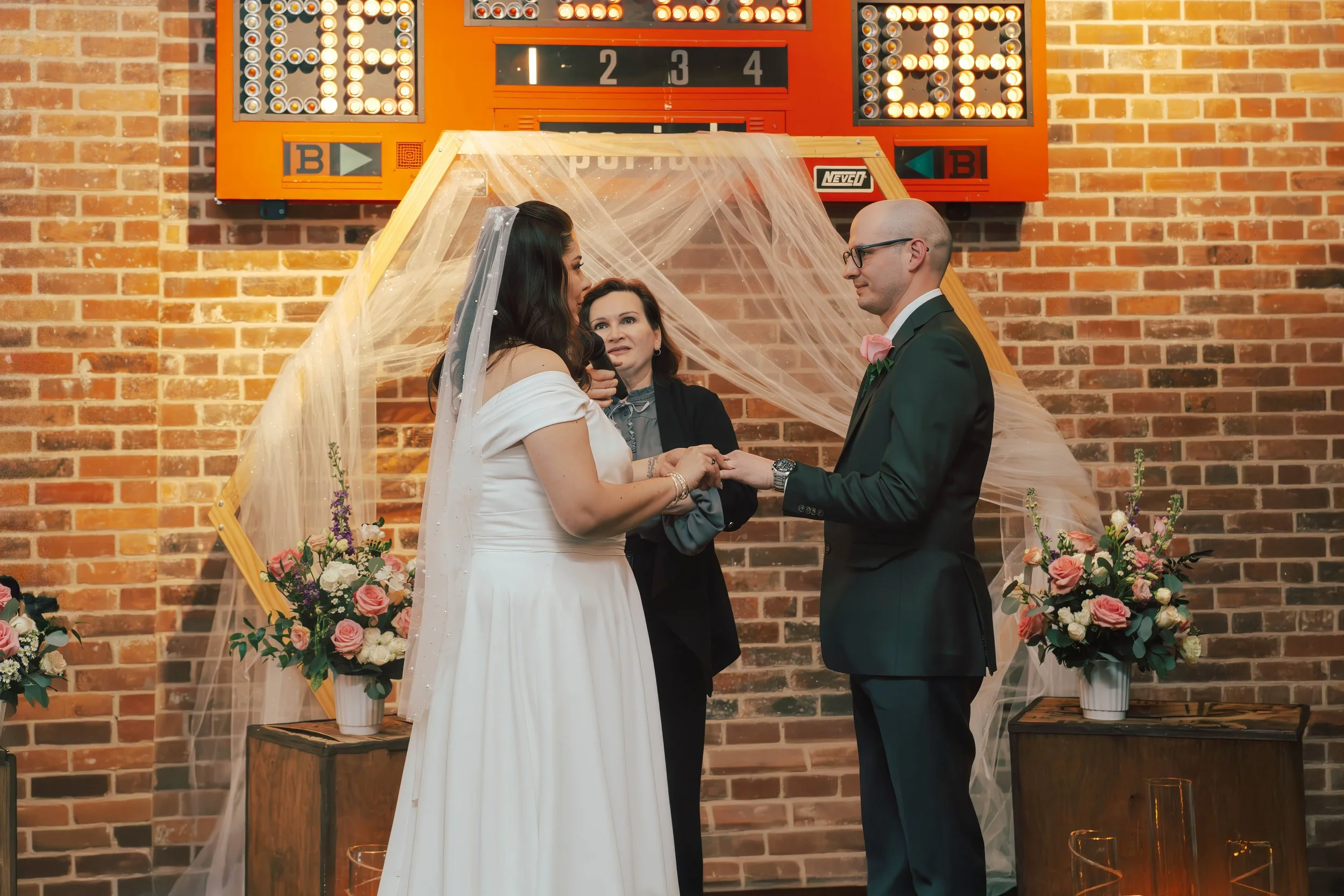 A bride and groom exchange vows during a wedding ceremony, holding hands, with a woman officiant in the background, in front of a brick wall decorated with flowers and a scoreboard-like backdrop.