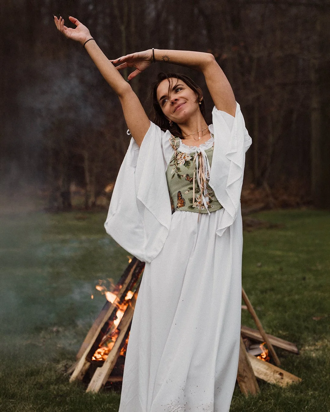 A woman with short dark hair, wearing a white dress with wide sleeves, is standing outdoors in front of a small bonfire. She is smiling and has her arms raised with hands positioned gracefully.