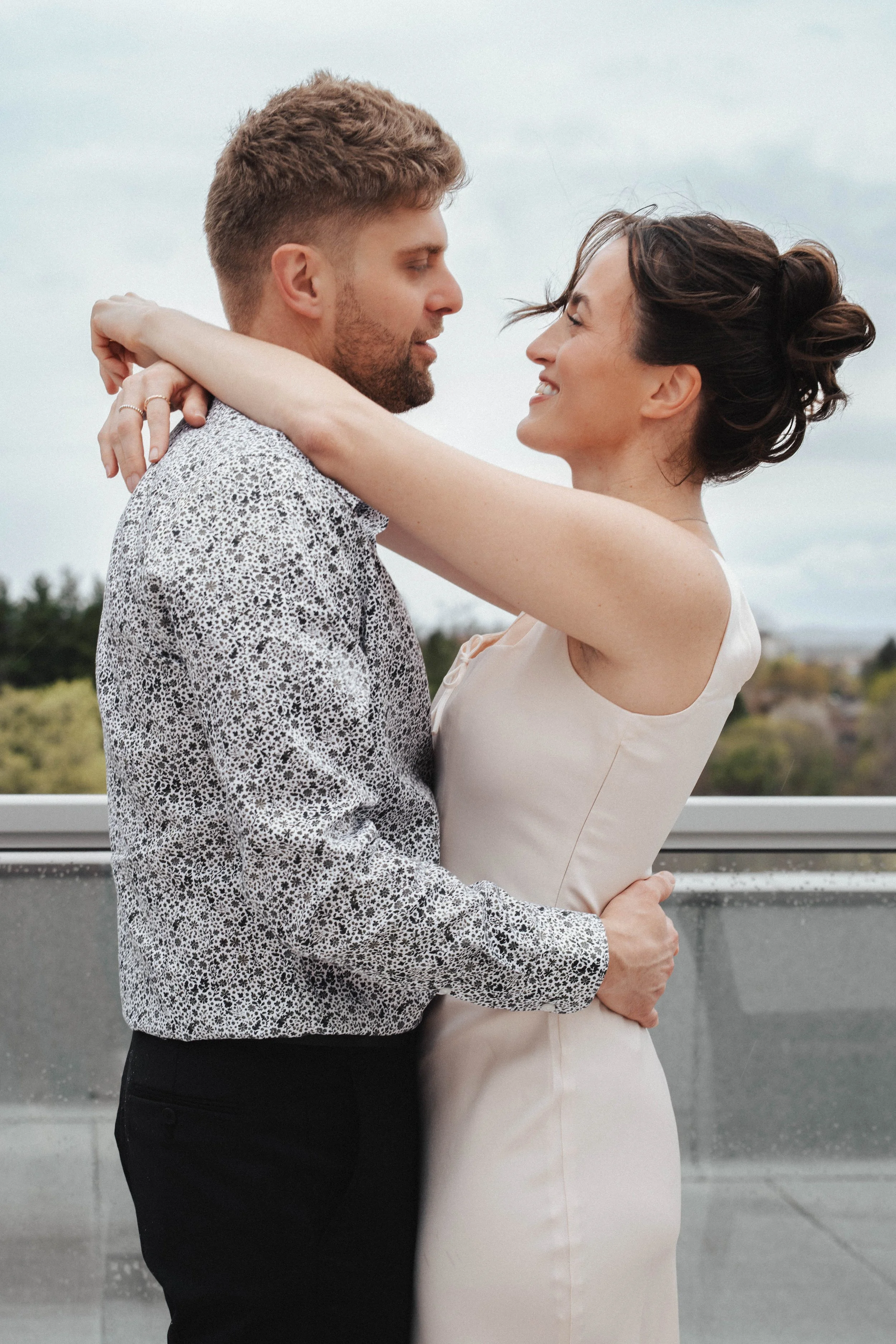 A couple embracing outdoors on a cloudy day. The man wears a patterned shirt, and the woman wears a light-colored dress. They look into each other's eyes, smiling, with arms wrapped around each other.