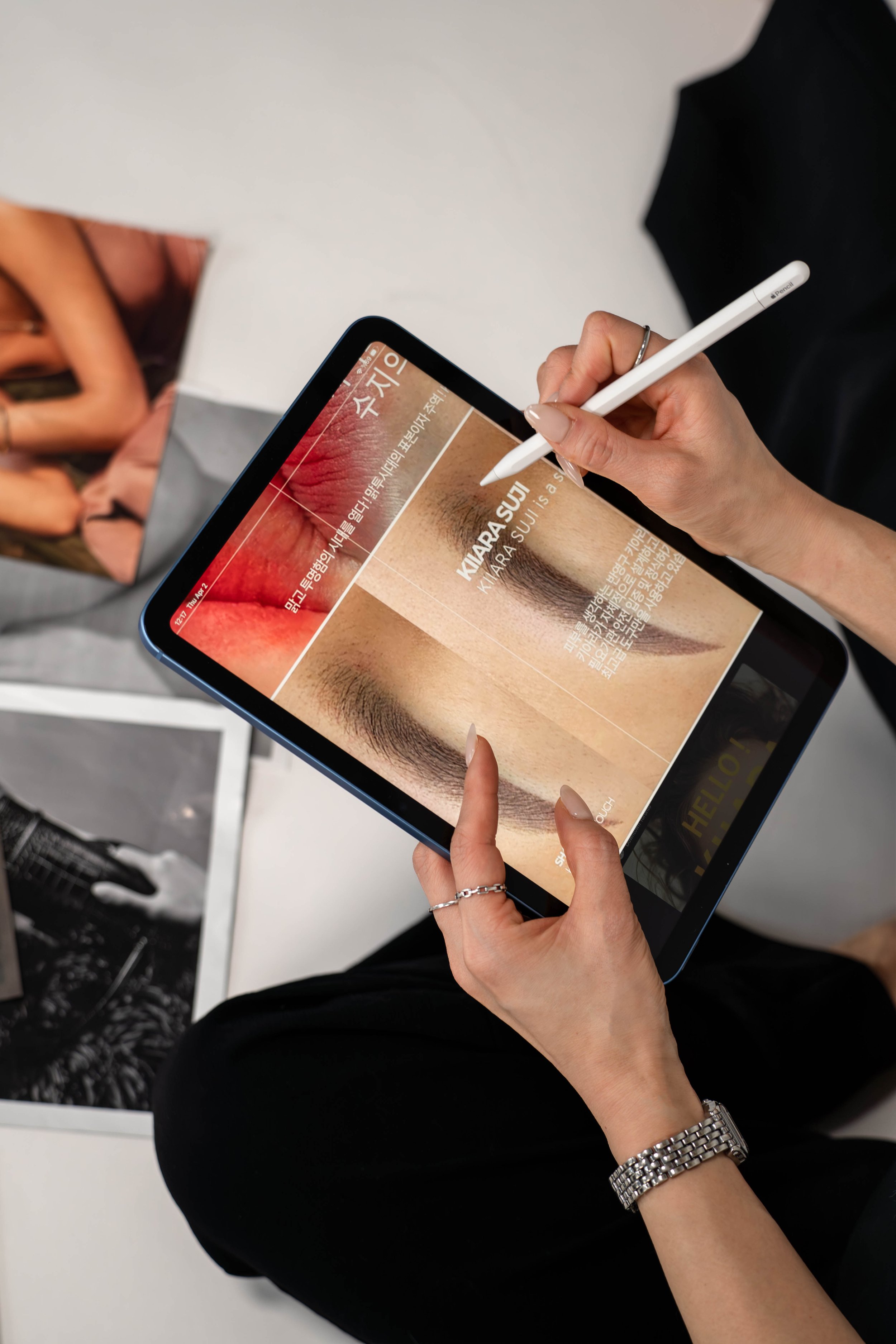 Person using a tablet with a stylus, viewing an article about eyebrow makeup or tattooing, with a close-up of a woman's eyebrow and lips on the screen.