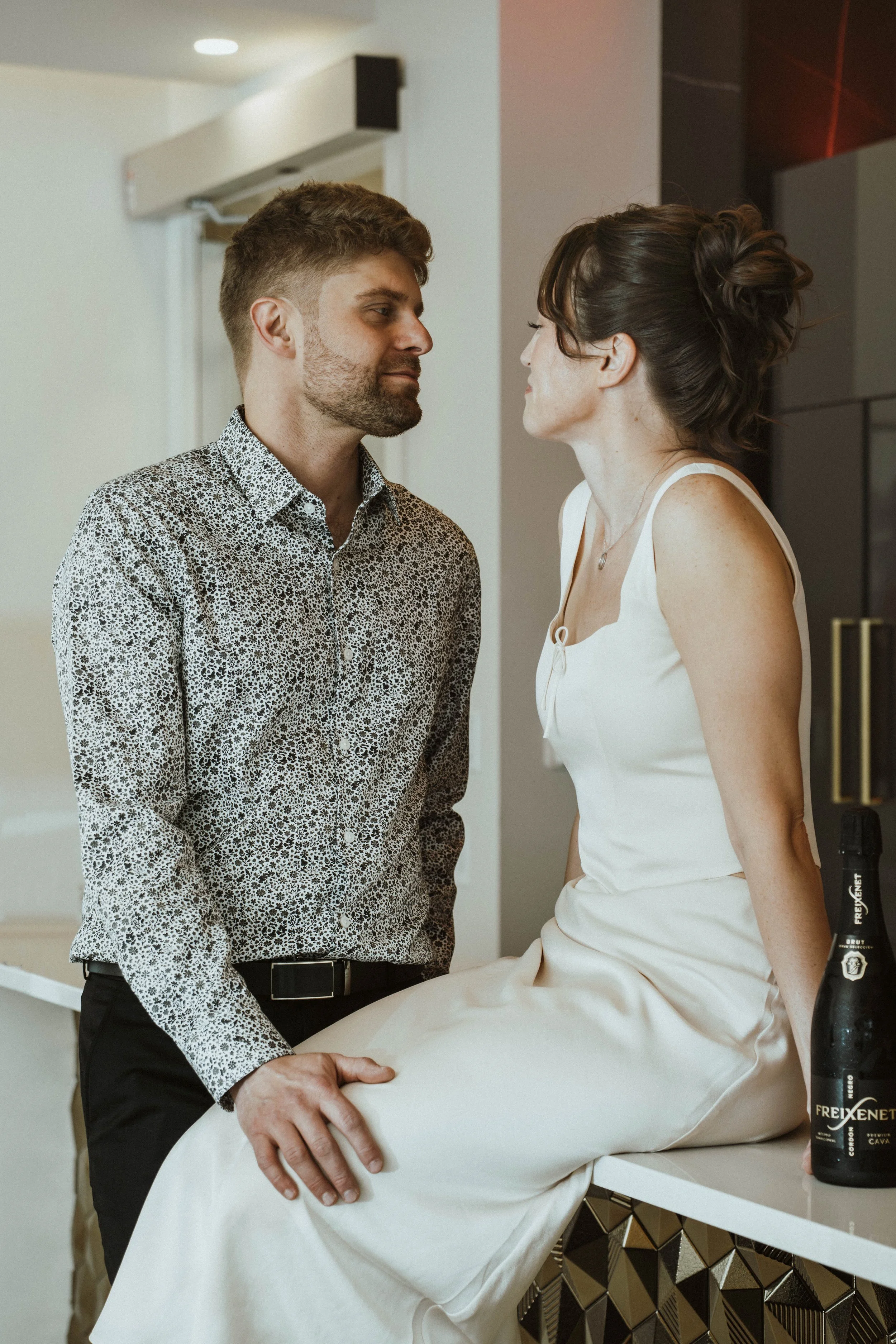 A man and woman face to face in a close, intimate moment in a modern interior space. The man is dressed in a patterned shirt and dark trousers, with his hand resting on a table. The woman is sitting on the table, wearing a white dress, with her hair 