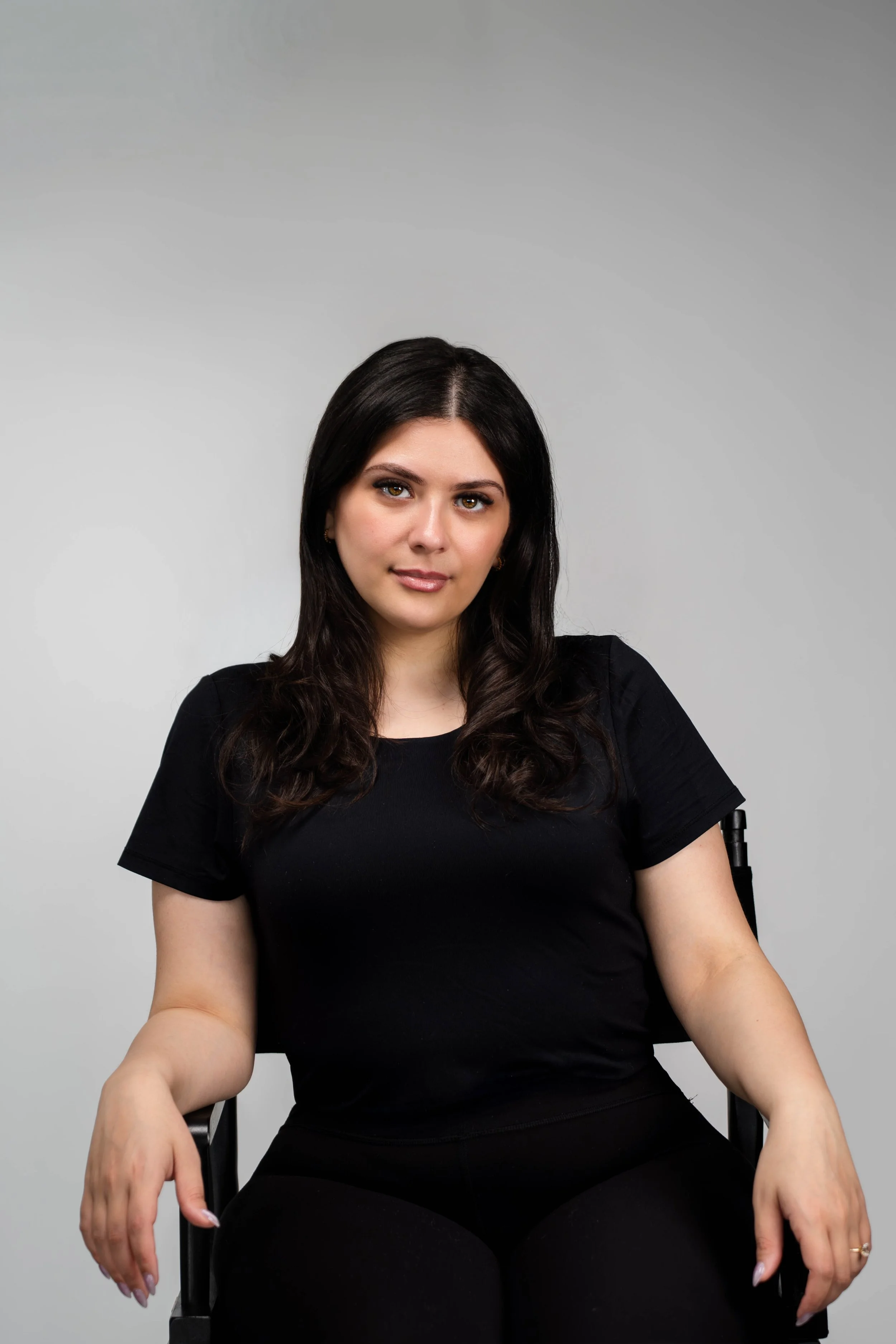 Portrait of a woman with long dark wavy hair, wearing a black t-shirt, seated against a plain gray background.