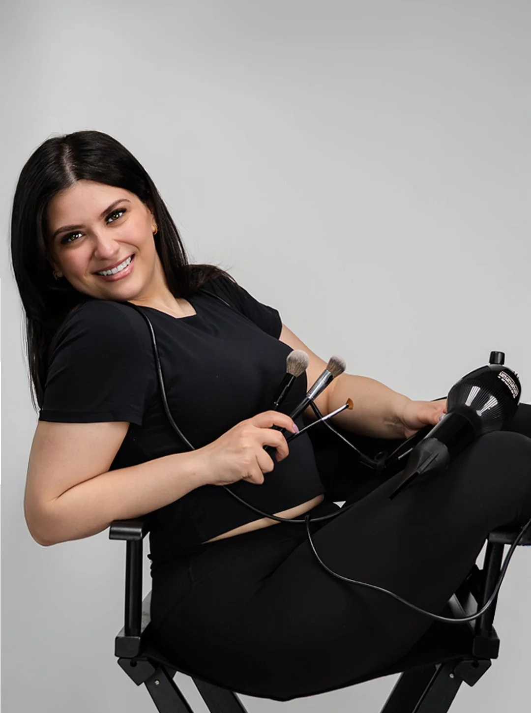 Woman dressed in black, sitting on a chair, smiling, holding makeup brushes and a hairdryer, with a professional makeup artist appearance.