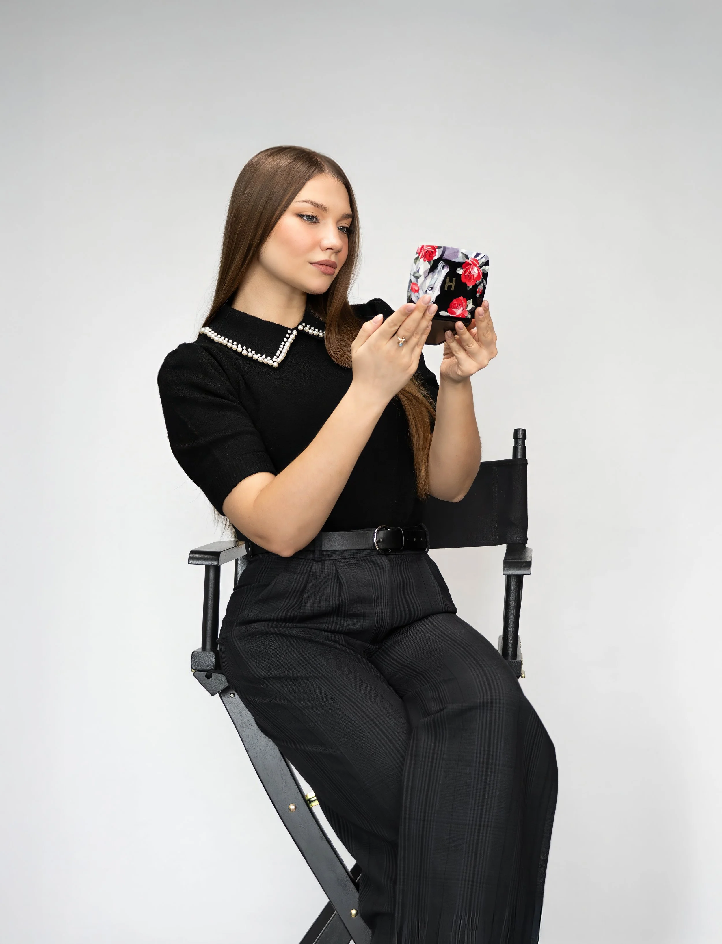 A young woman with long brown hair sitting on a director's chair, holding and looking at a decorative box with floral and face designs.