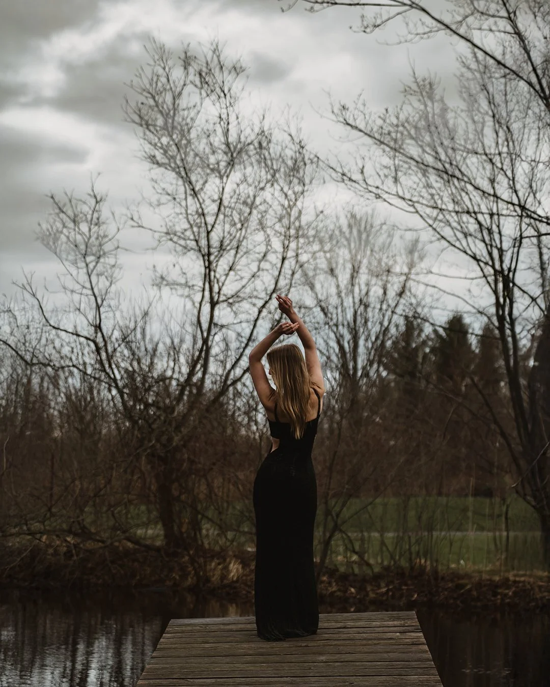 A woman standing on a wooden dock near water, with bare trees in the background, wearing a long black dress, raising her arms above her head, under a cloudy sky.