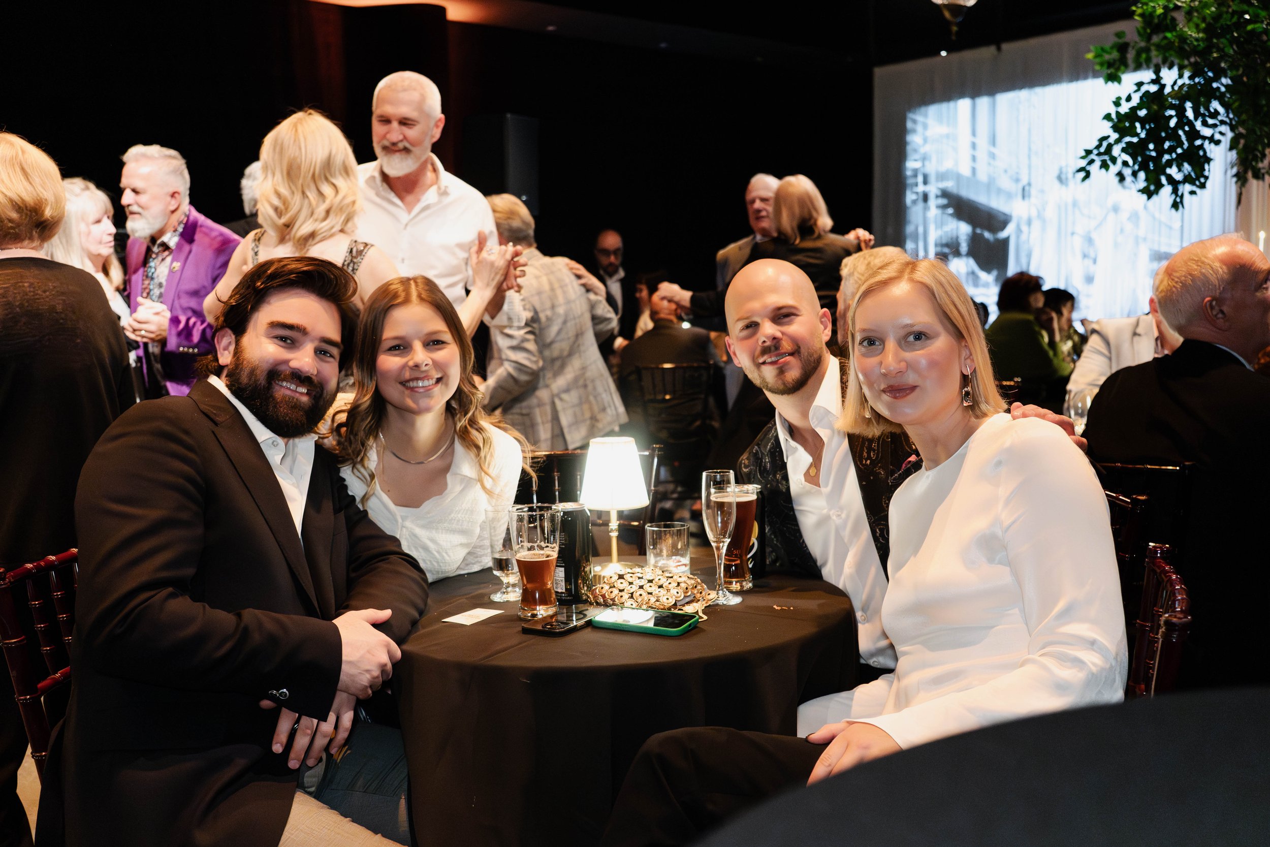 Four people sitting at a round table enjoying a social event, with many people dancing and talking in the background.