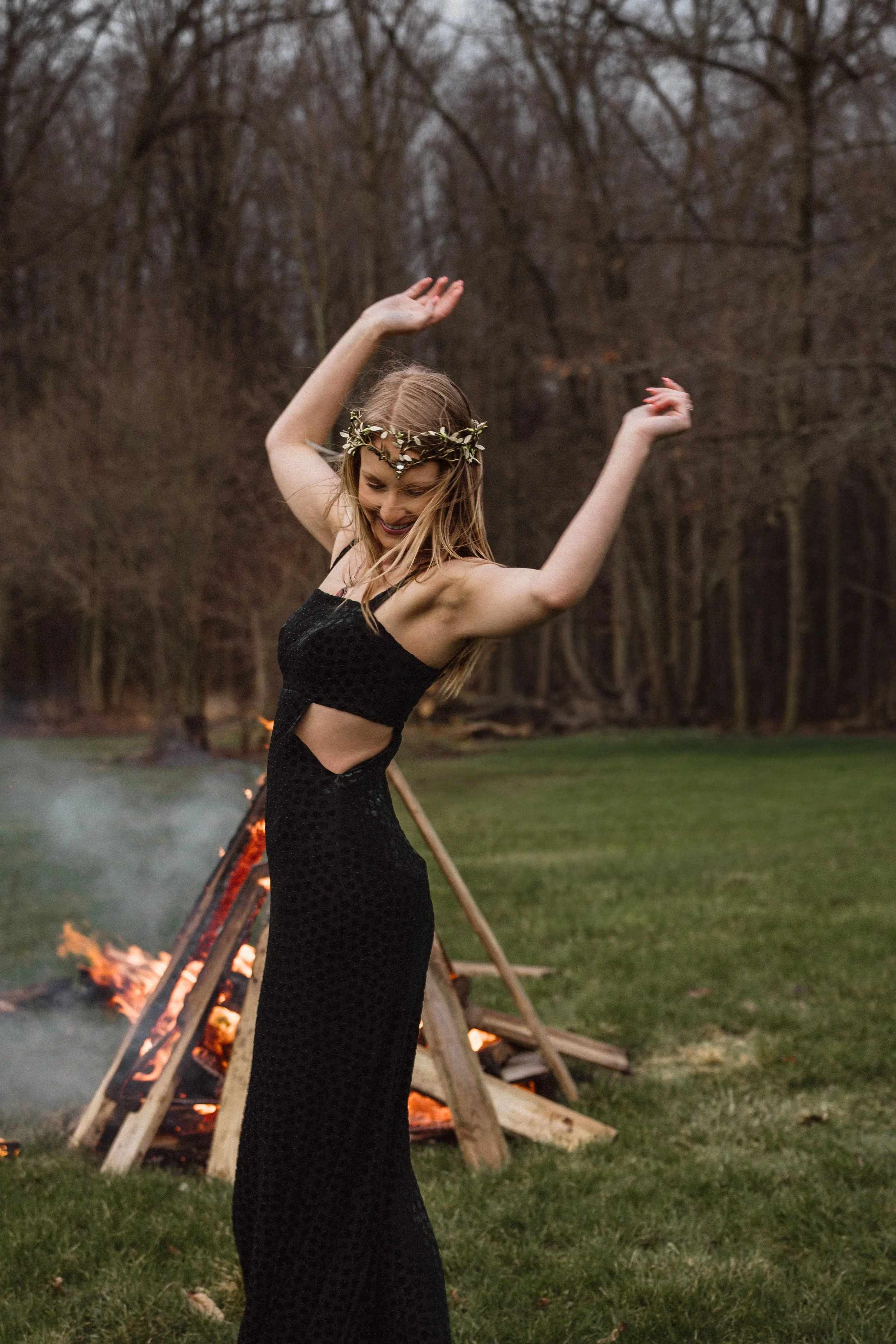 A young woman in a black dress with cutouts dancing outdoors near a bonfire in a forested area.