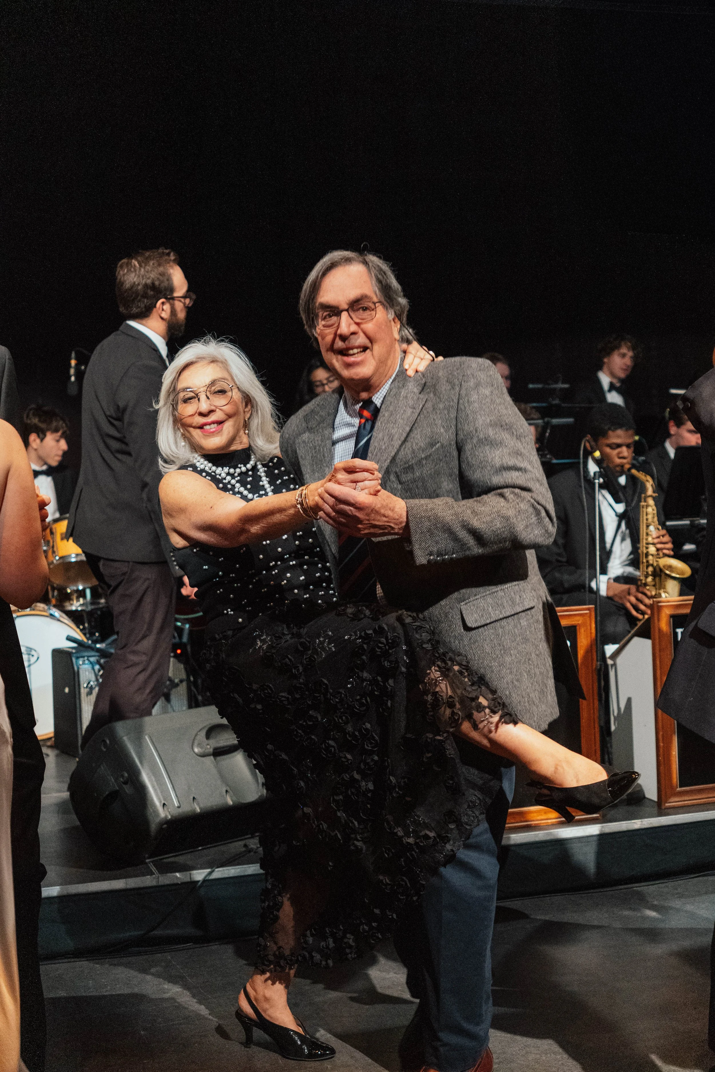 A woman in a black dress and heels is being lifted by a man in a gray suit on a stage with a band and orchestra in the background.