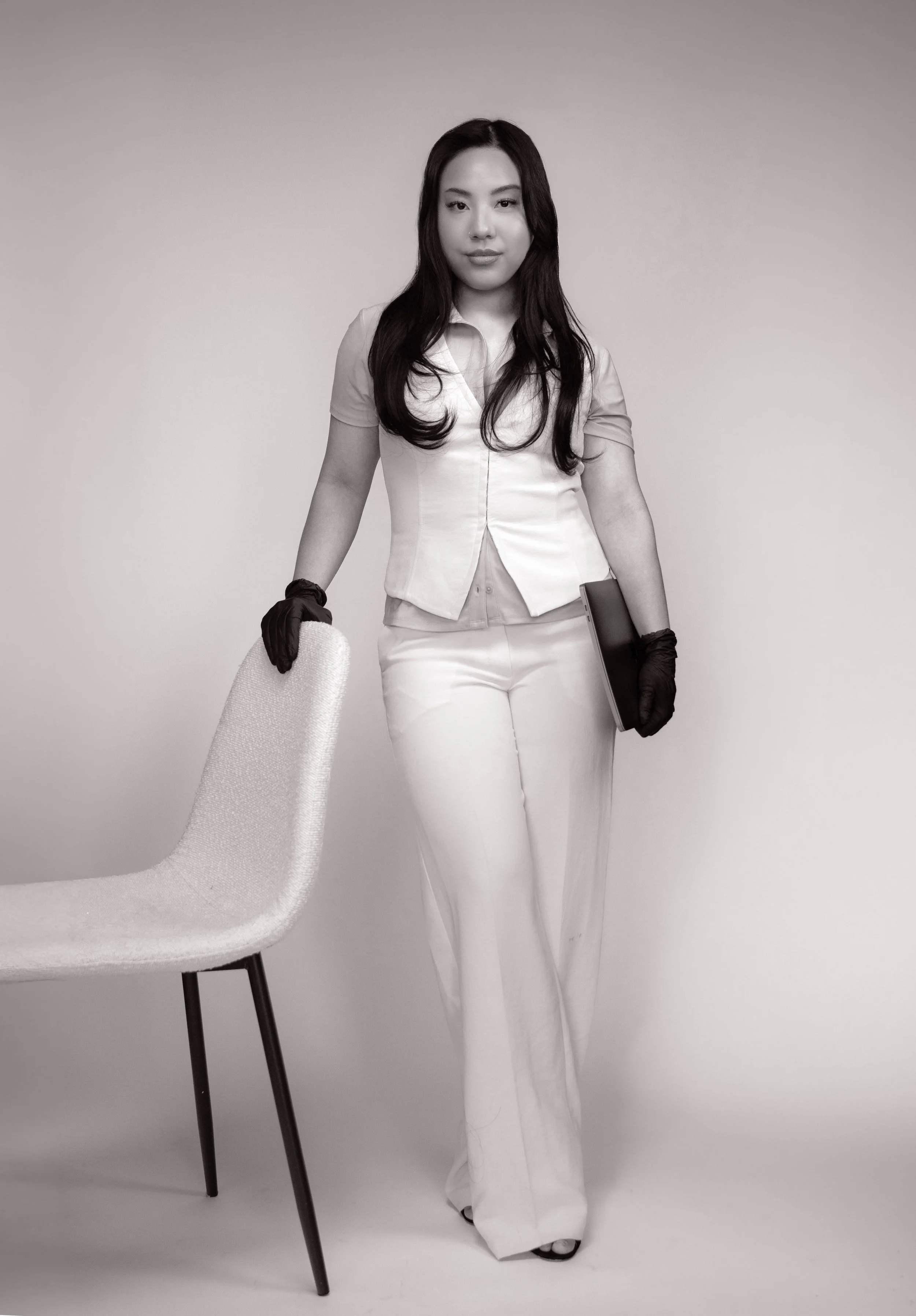 A woman standing next to a chair, wearing a white suit with black gloves and holding a laptop and a closed gaming device, in front of a plain background.