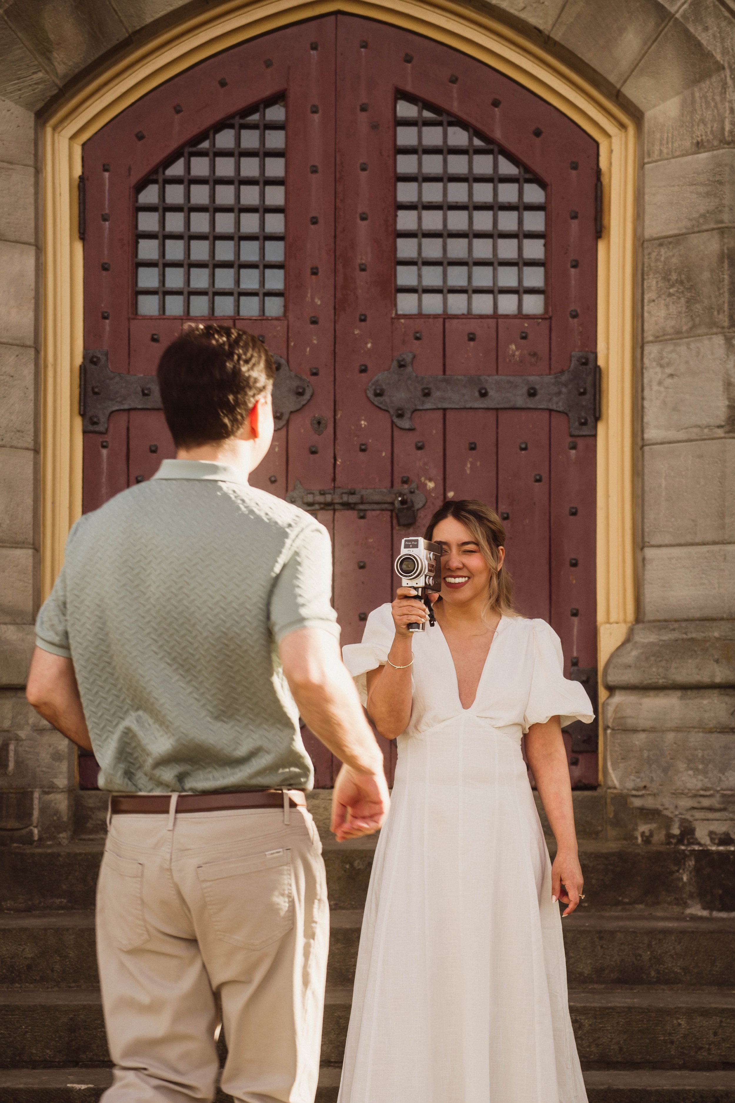 A woman in a white dress smiles happily while holding a gun, standing in front of a large wooden and metal door with a man in a light-colored shirt and beige pants standing nearby.