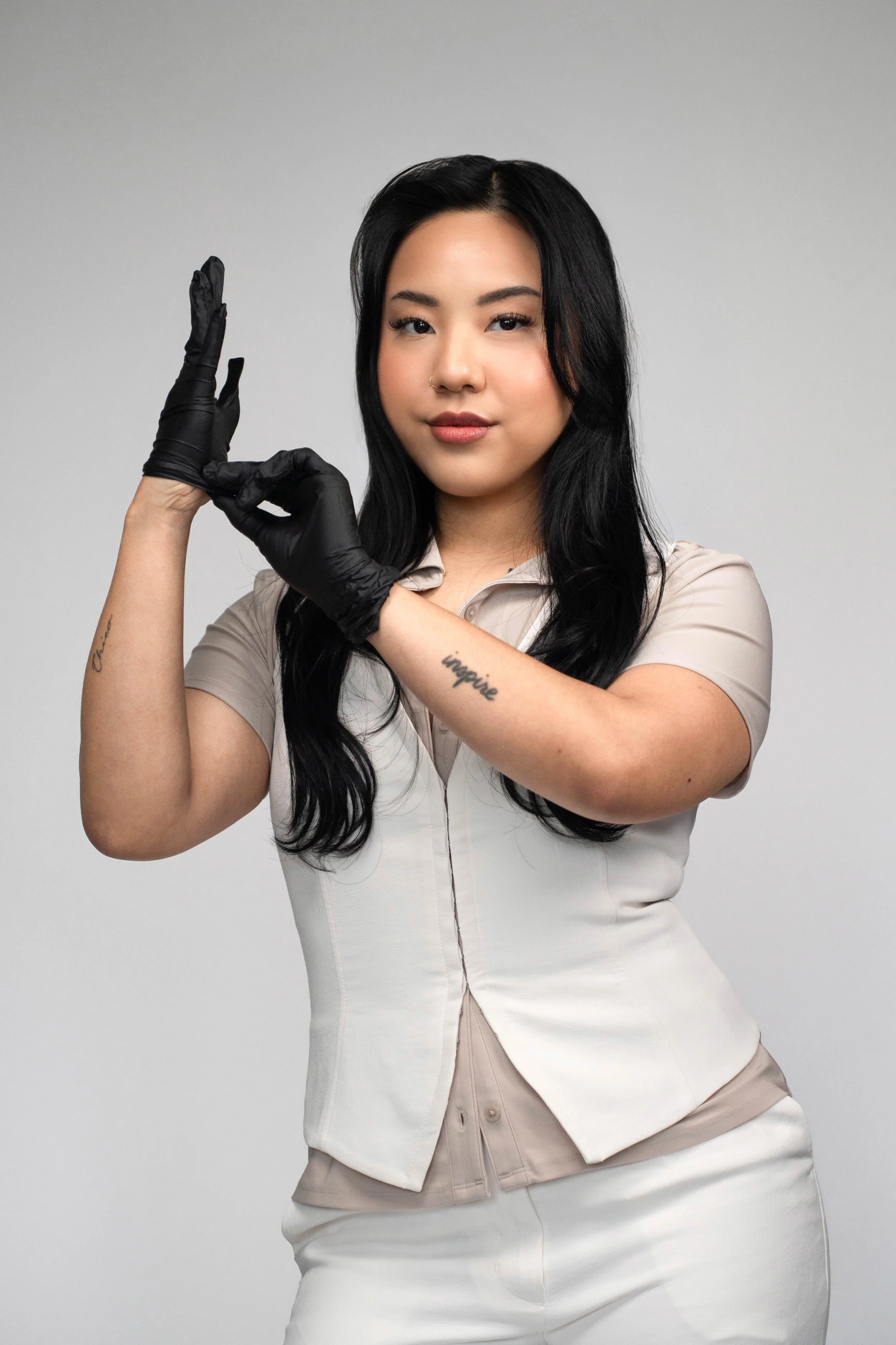 A young woman with long black hair, wearing a beige shirt and gloves, making an X sign with her hands.
