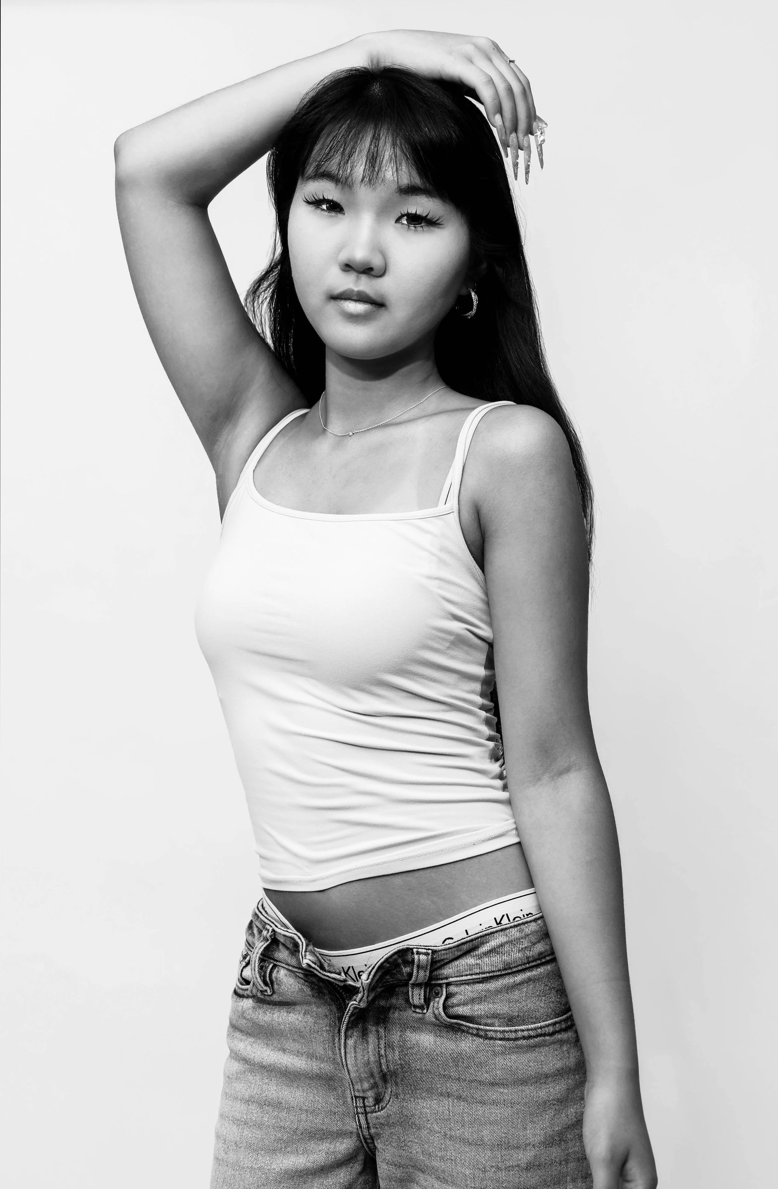 Black and white photo of a young woman with long dark hair, wearing a tank top and jeans, posing with one arm raised and hand resting on her head.