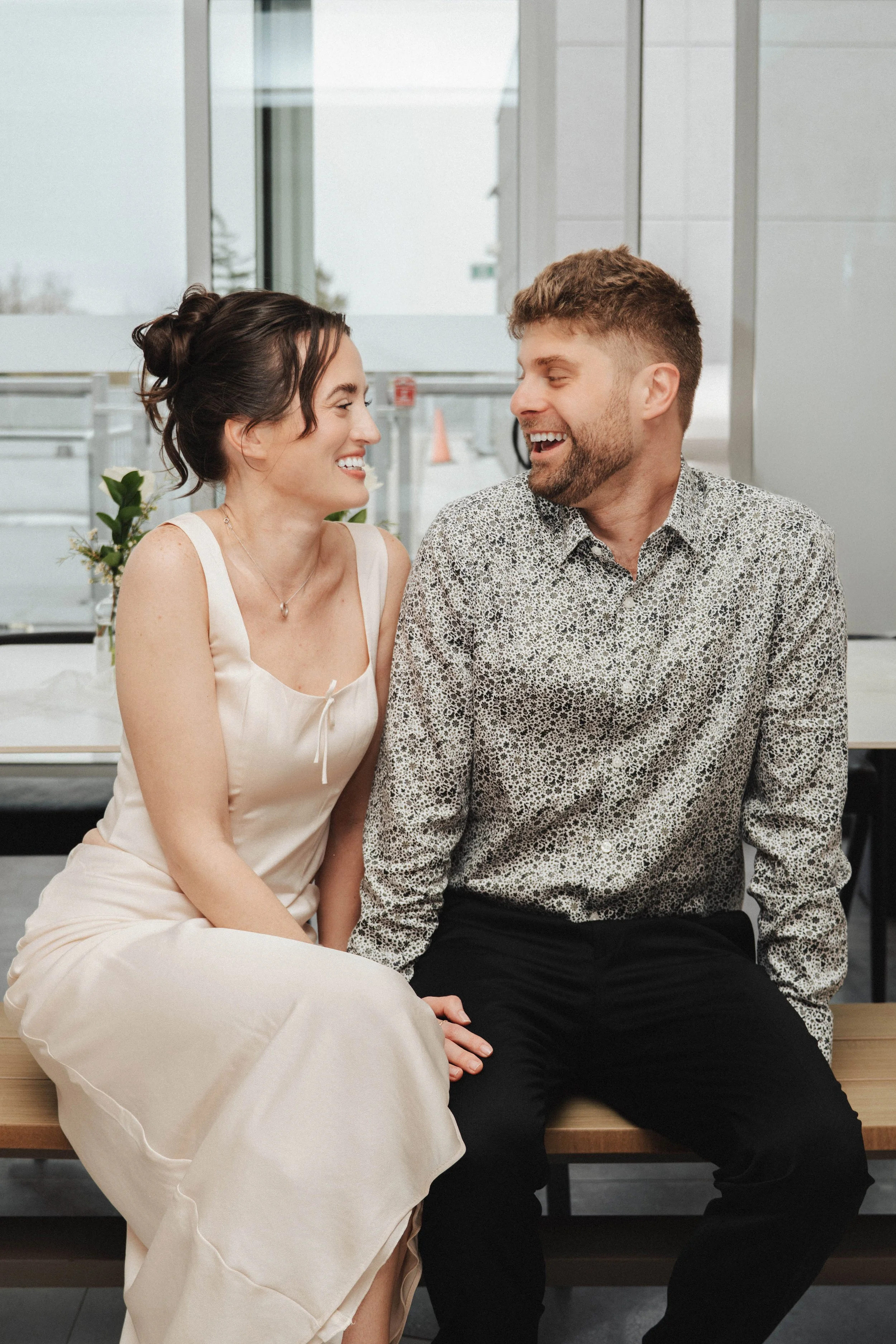 A woman and a man are sitting on a bench inside a building, smiling at each other and making eye contact. The woman is wearing a light-colored dress, and the man is wearing a patterned shirt and black pants. They appear to be happy and engaged in con