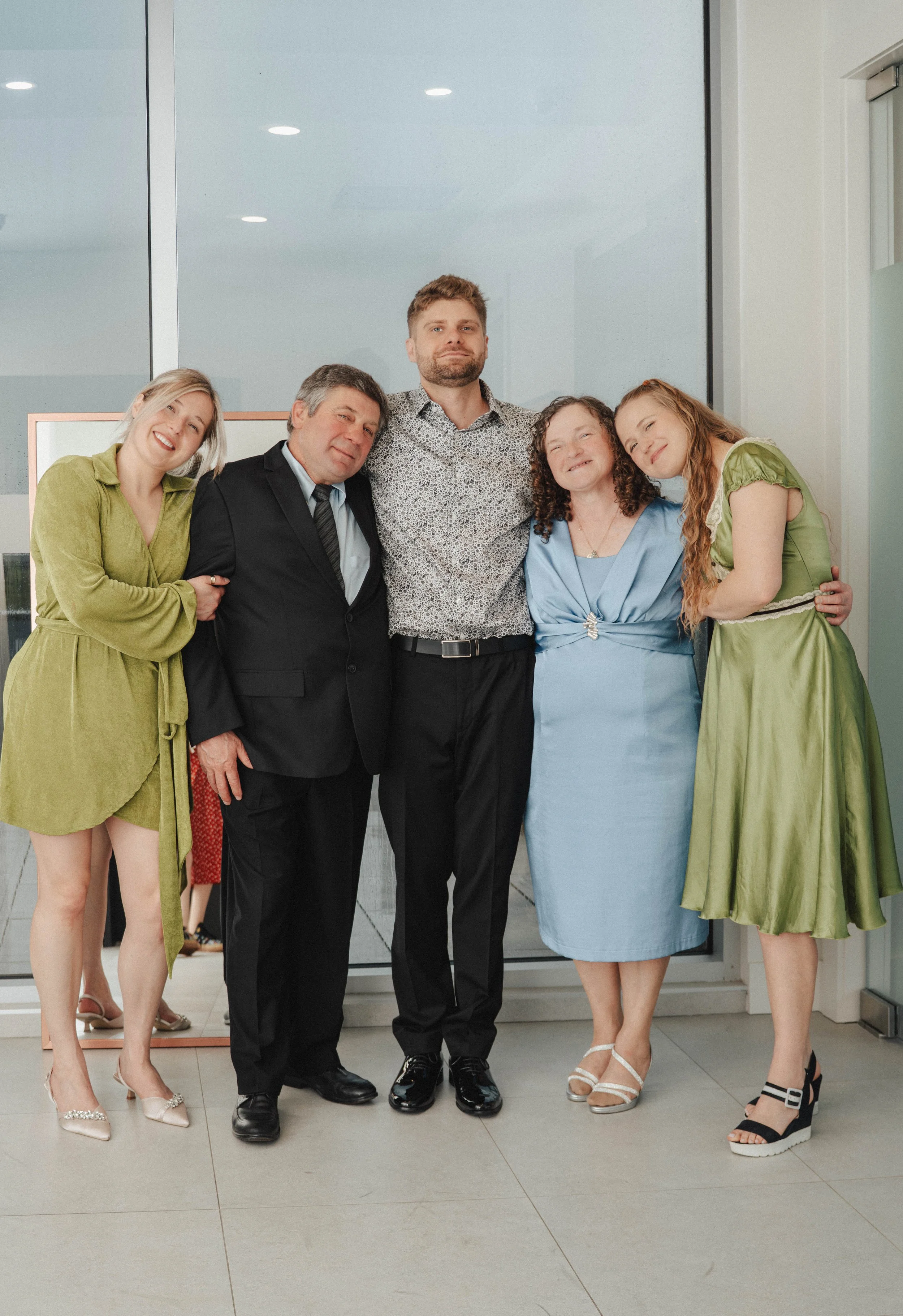 Group of five people standing in front of a glass door, dressed in semi-formal attire, smiling, with a woman and a man visible in the background.