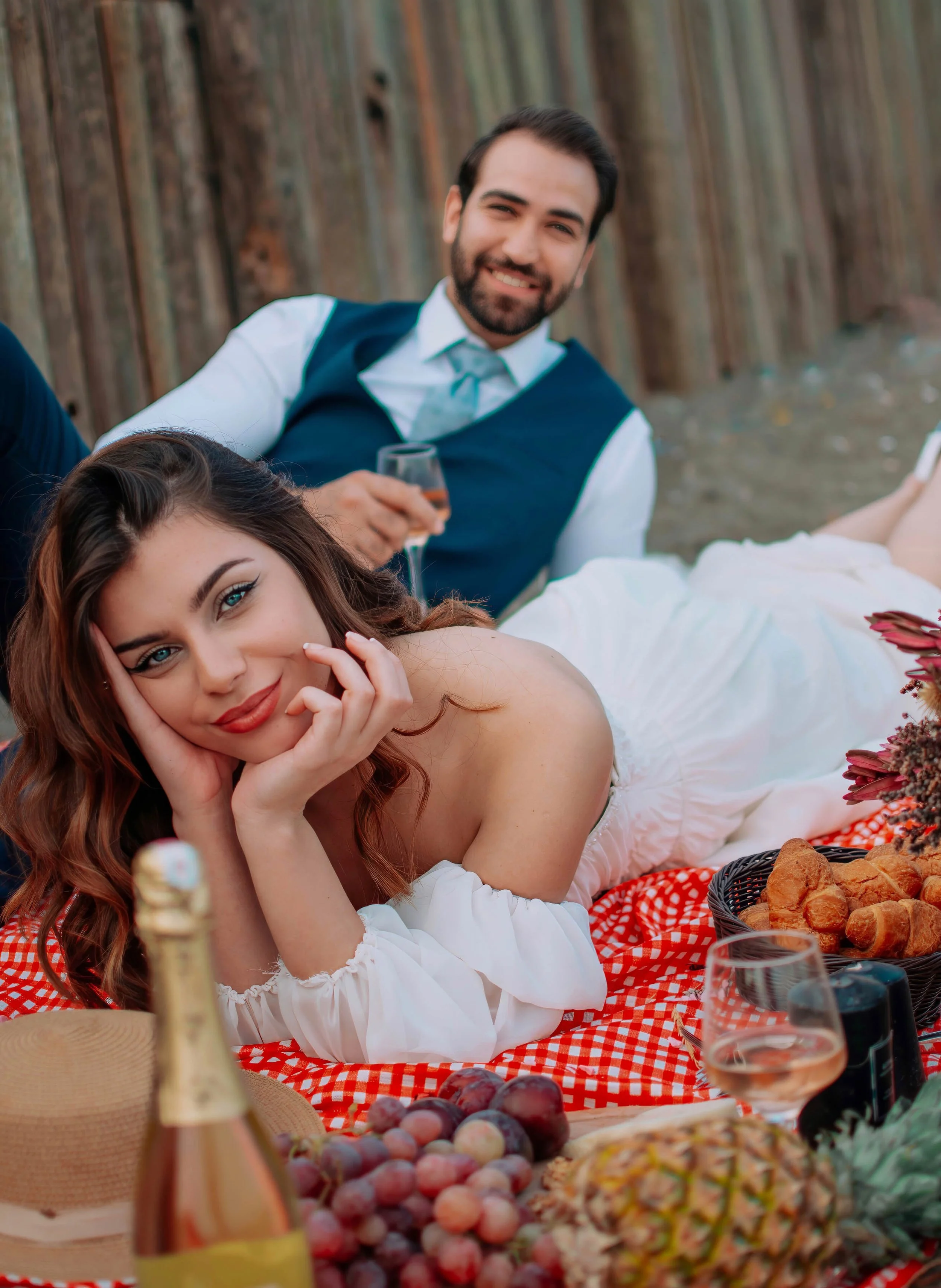 A woman with long brown hair lies on her side on a picnic blanket, resting her head on her hand and smiling at the camera. A man in a white shirt and blue vest sits behind her, holding a glass of wine and smiling. The picnic table is decorated with f