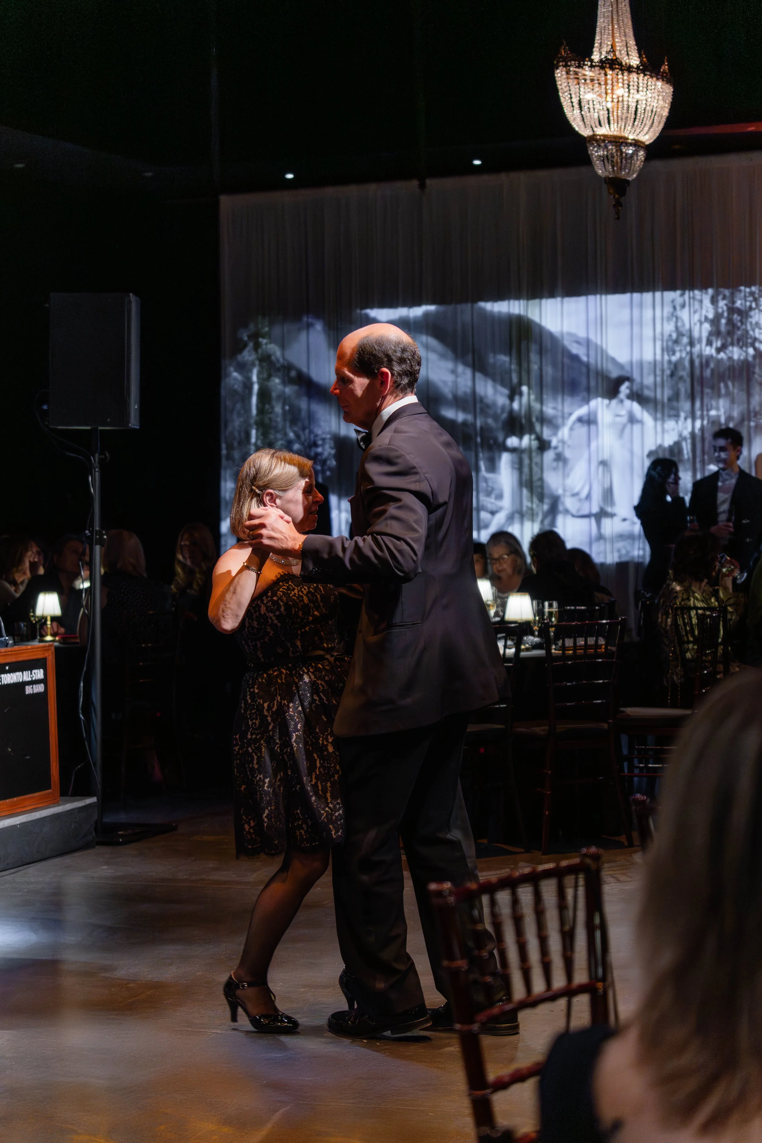 A man and woman dancing closely at a formal event with a black and white photo projected on a curtain behind them. Guests sit at tables around them, and a chandelier hangs from the ceiling.