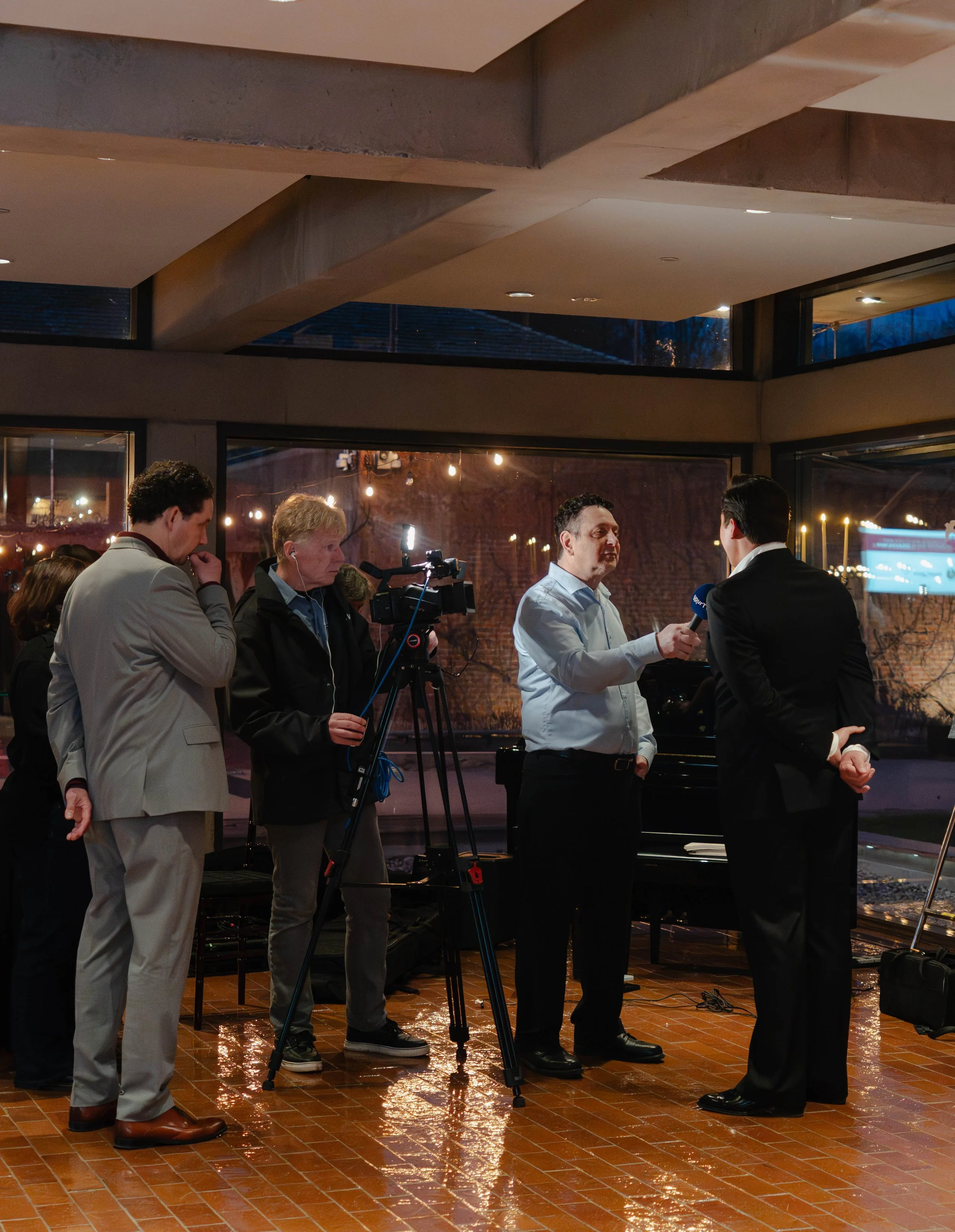 A man being interviewed by a reporter on a video camera setup. Several people observing, some with hand on face, indoors in a well-lit room with large windows showing a nighttime cityscape outside.