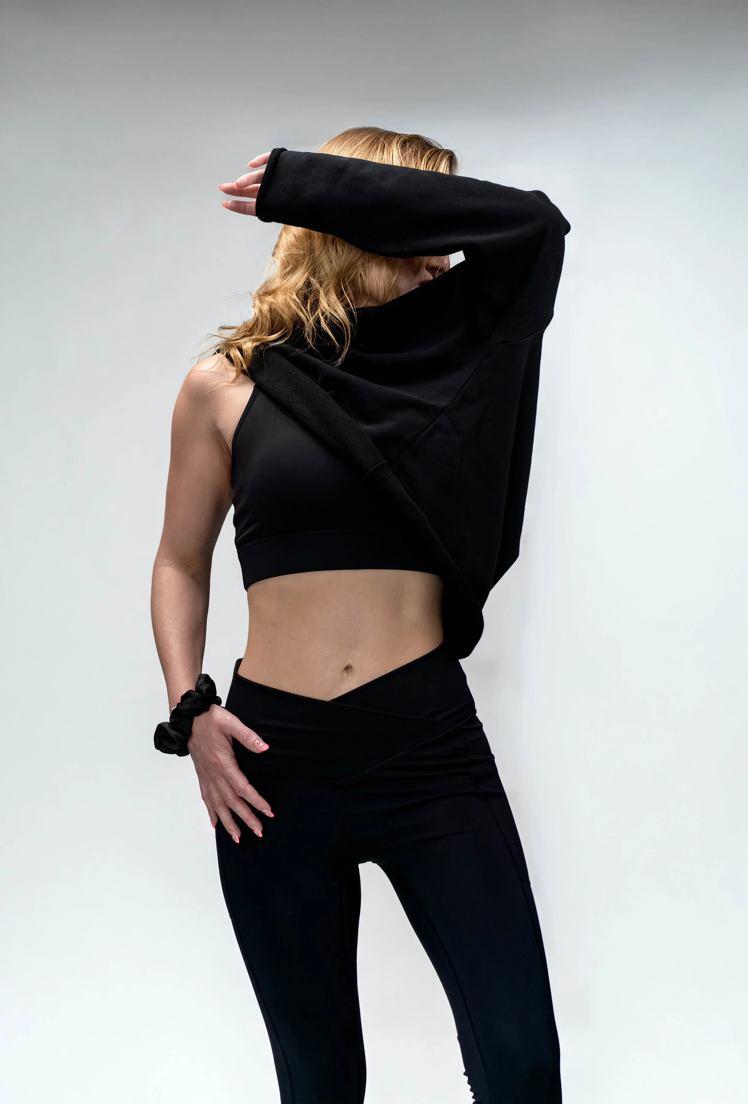 A woman with curly blonde hair poses with her right arm covering her face, wearing a black sports bra, high-waisted black leggings, a black oversized sweatshirt draped off her shoulder, and a black scrunchie on her left wrist against a plain white ba