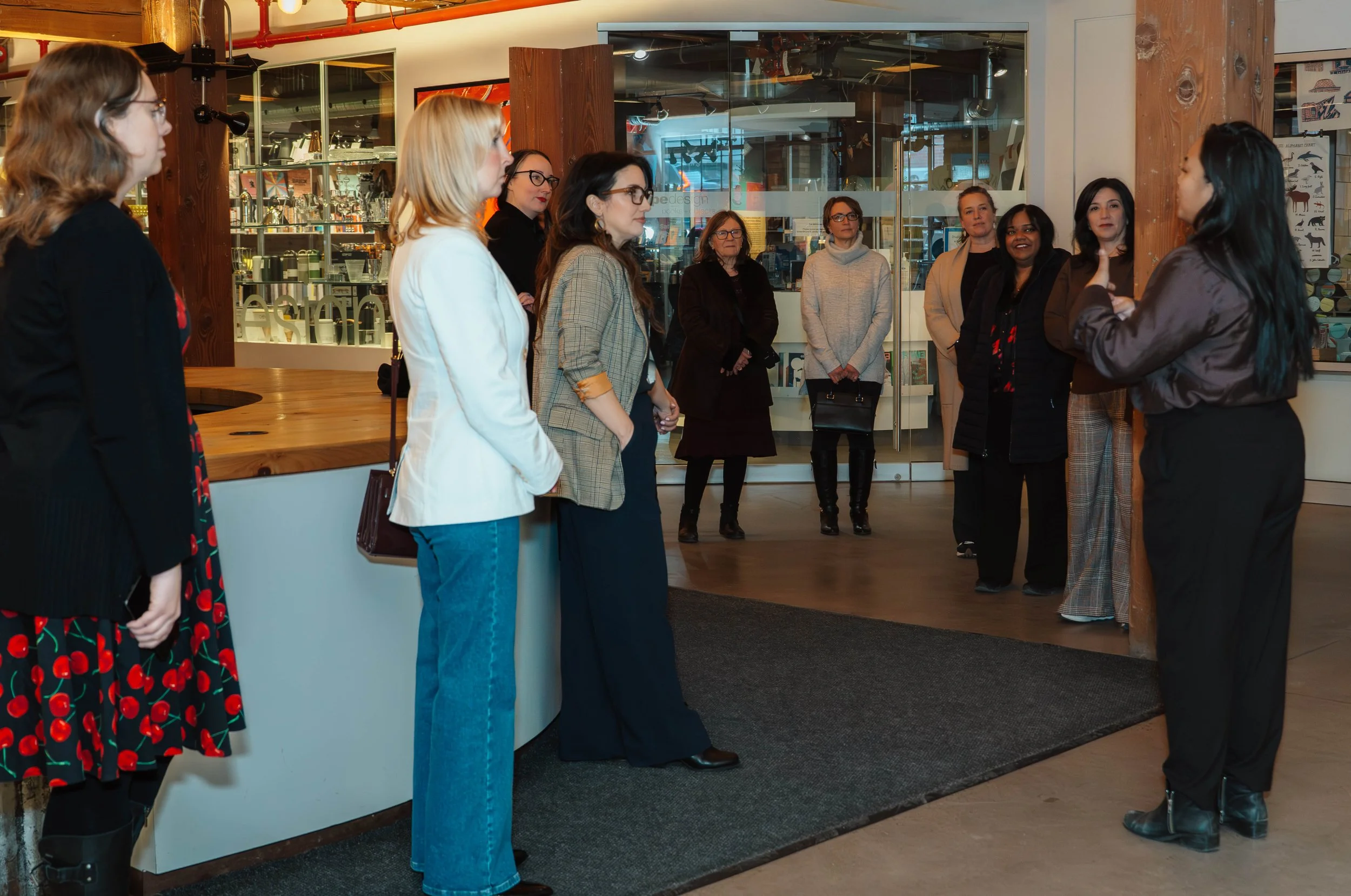 A woman is giving a tour or presentation to a group of women inside a modern, well-lit space, possibly a retail or office environment.