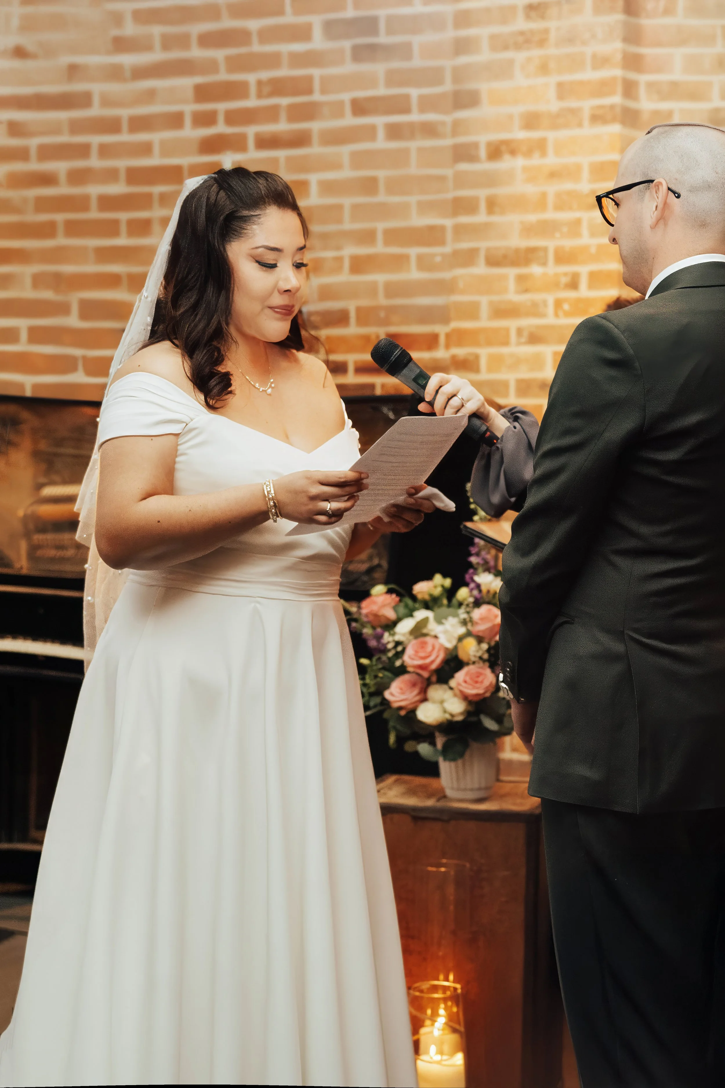 A bride and groom exchanging vows during their wedding ceremony indoors, with a brick wall background and floral arrangements.