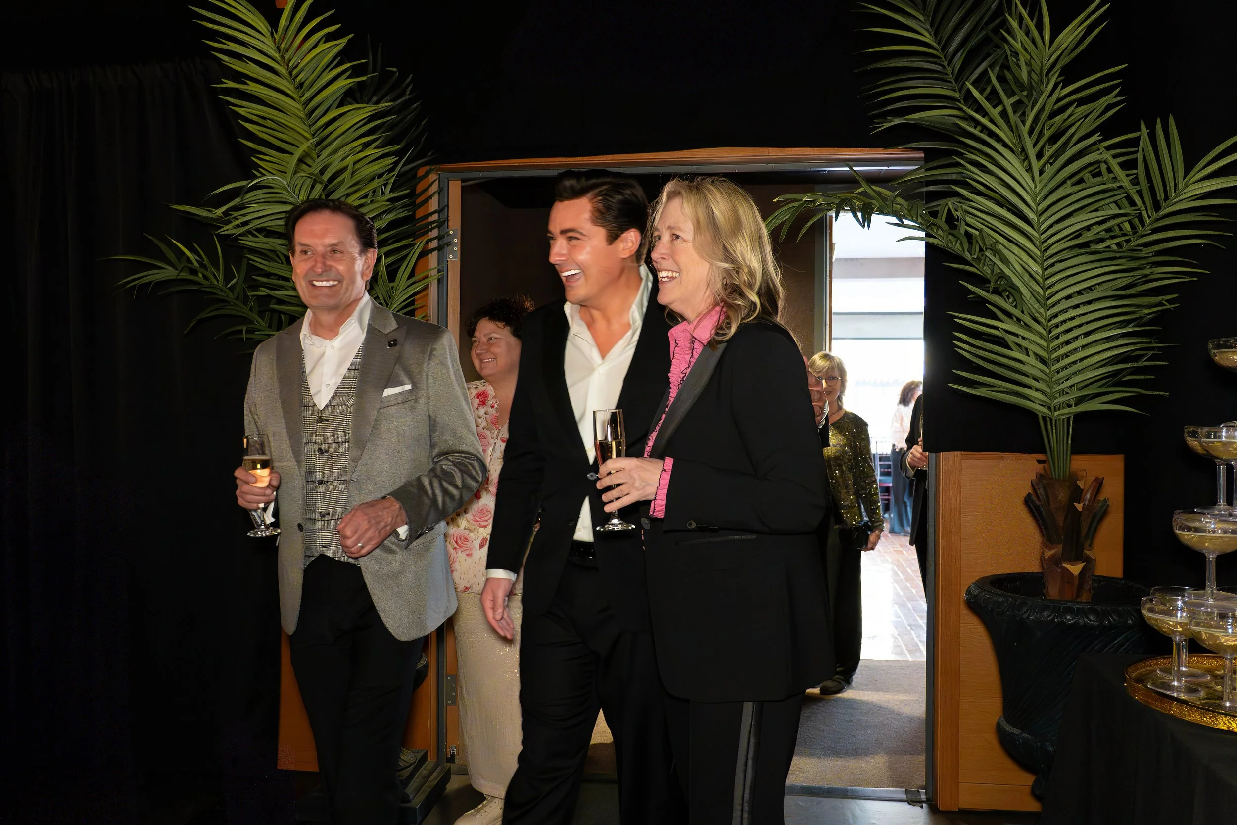 People at a social gathering, dressed in formal attire, holding glasses of wine, and smiling.