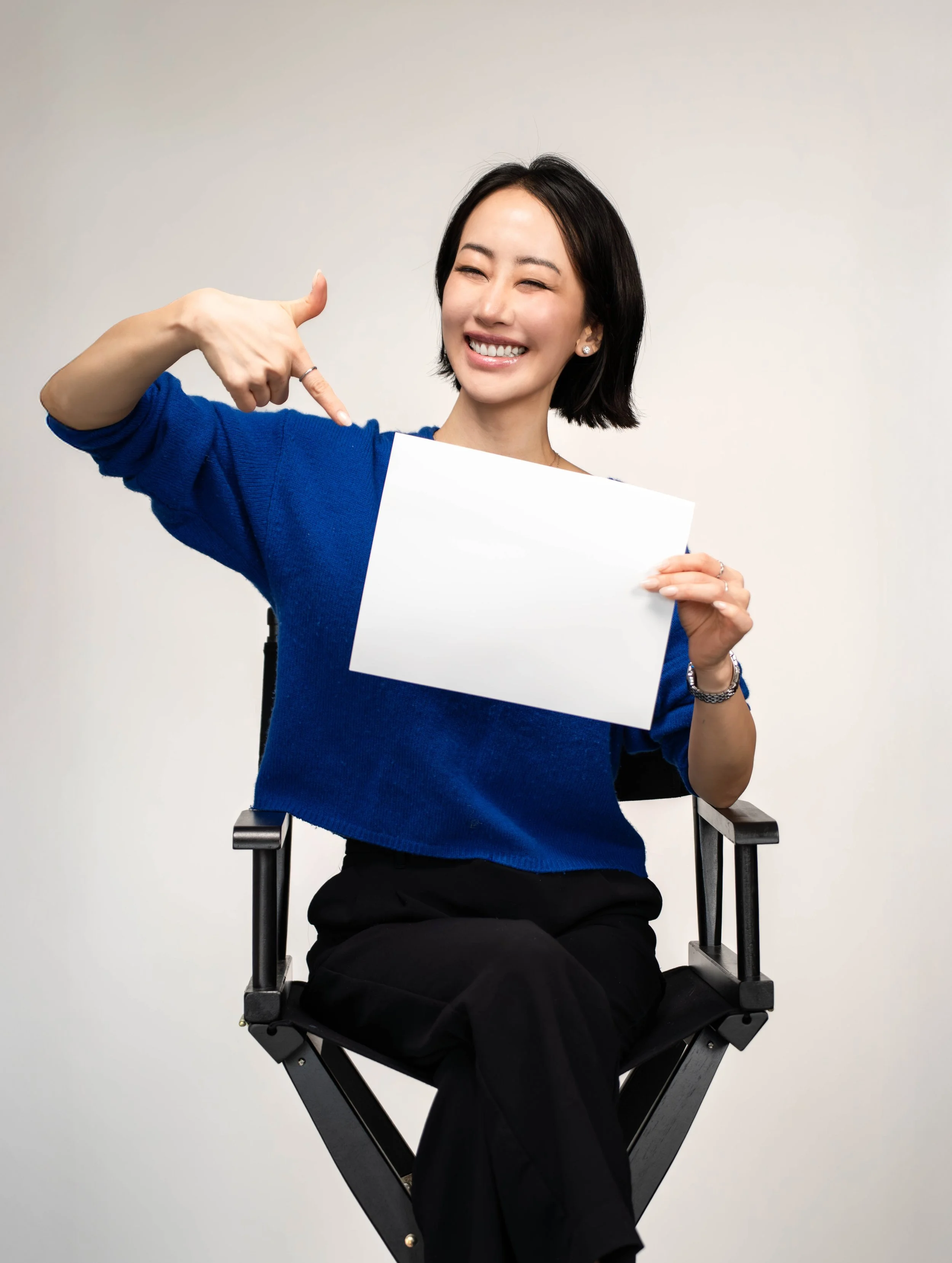 A woman smiling and pointing at a blank white sign she is holding, sitting on a director's chair against a plain white background.