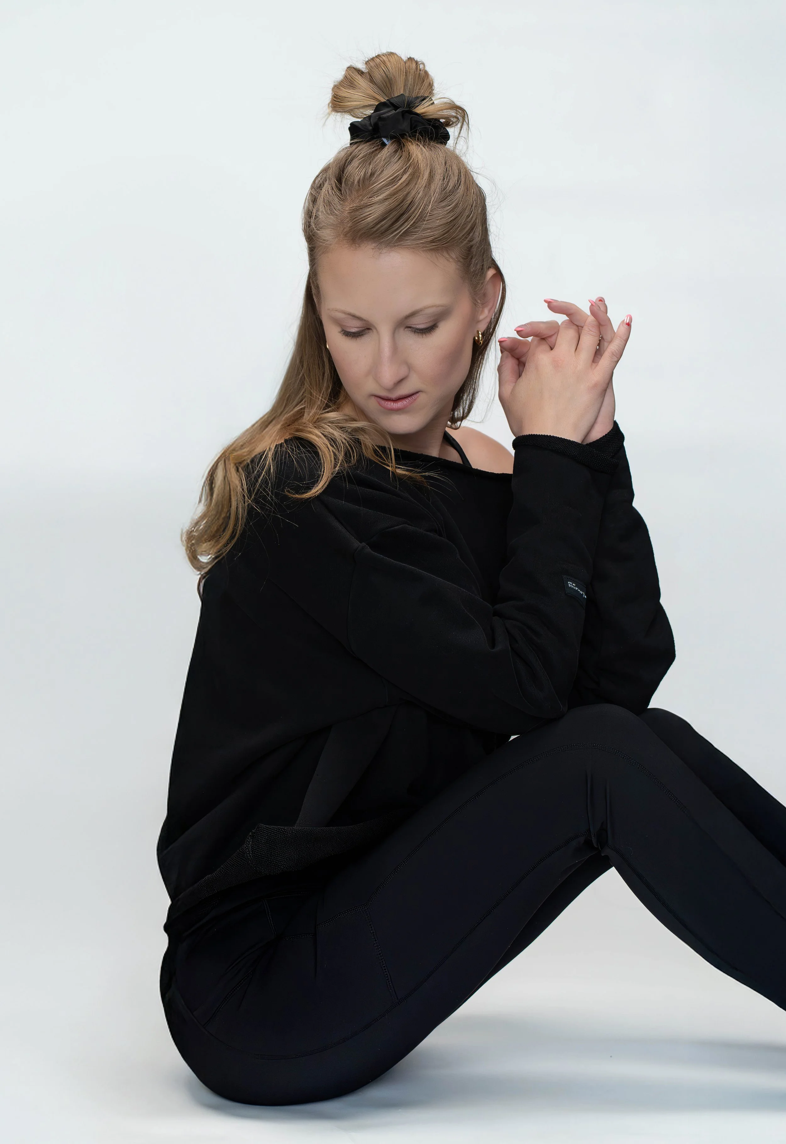 A woman with long, blonde hair tied up in a bun with a black scrunchie, sitting against a plain light background. She is dressed in black, wearing a black long-sleeved top and black pants, with her eyes closed and head tilted slightly downward, holdi
