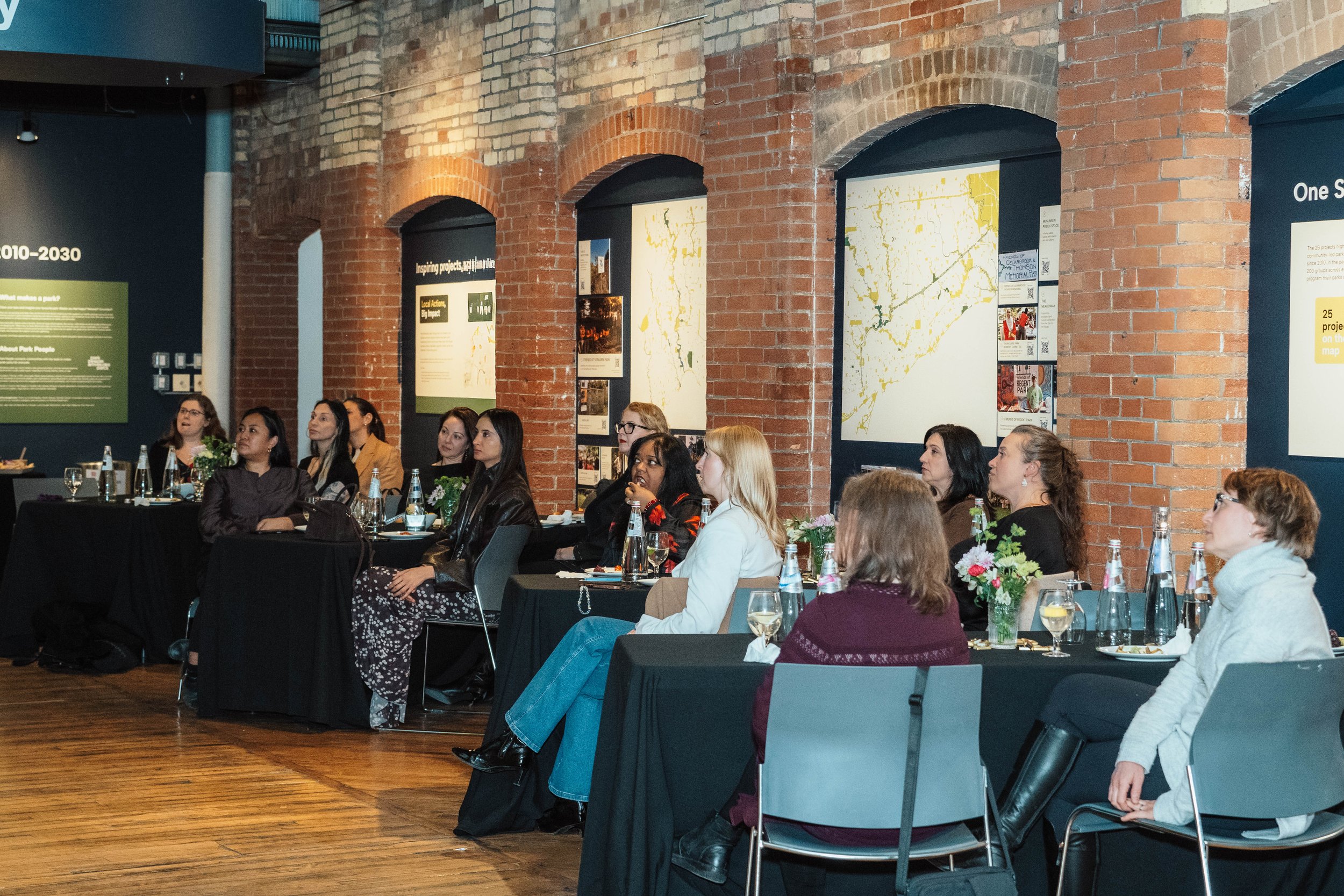 A group of women seated at tables in a conference room, attentively listening to a presentation. The room has brick walls and displayed posters on the walls.
