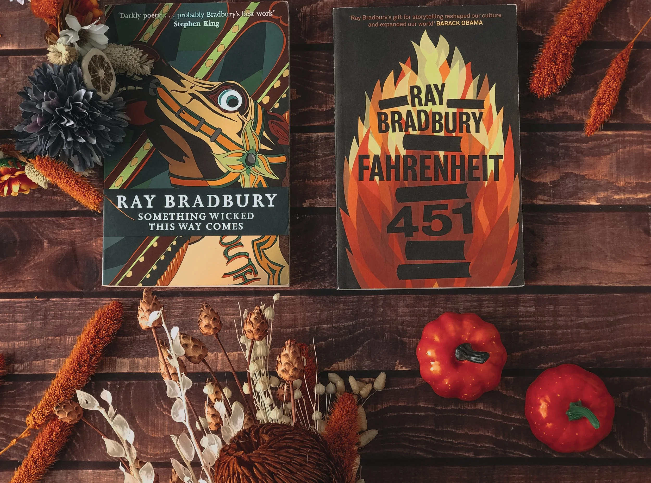 Two books by Ray Bradbury, one titled 'Something Wicked This Way Comes' and the other 'Fahrenheit 451', are placed on a wooden table. Around them are red mini pumpkins, dried flowers, and artificial orange and dark purple flowers for decoration.
