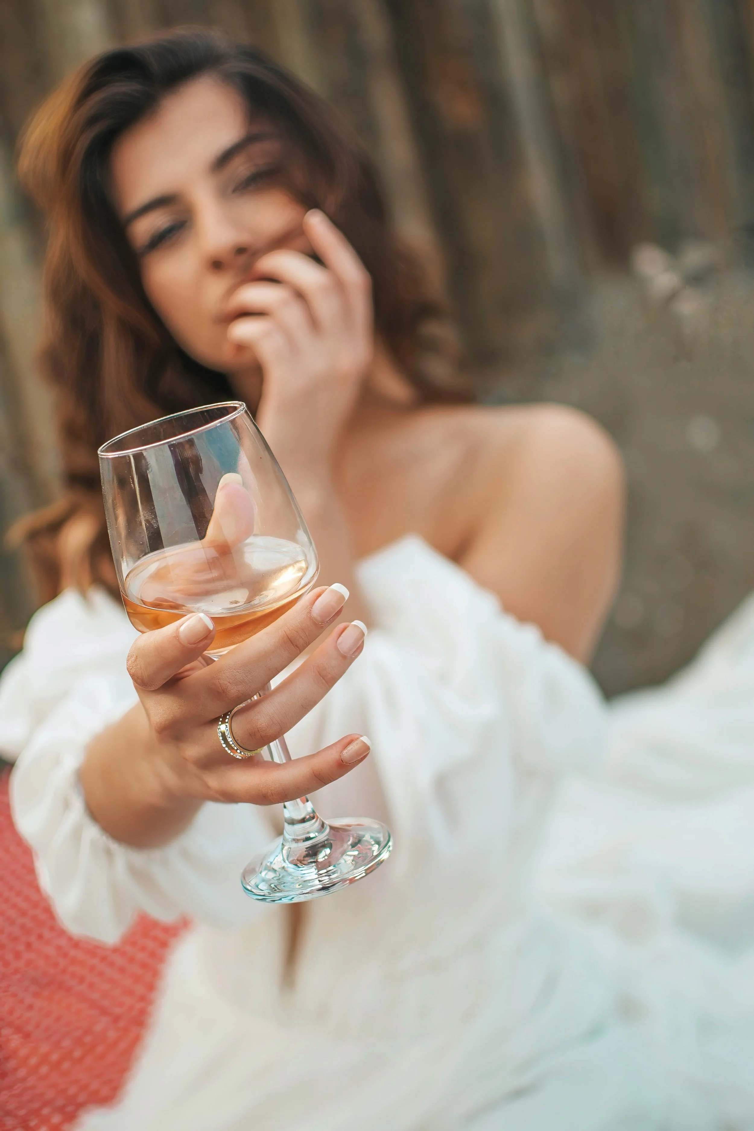 A woman with wavy brown hair holding a glass of rosé wine, partially covering her face with her hand, wearing a white off-shoulder top, with a blurred background of trees.