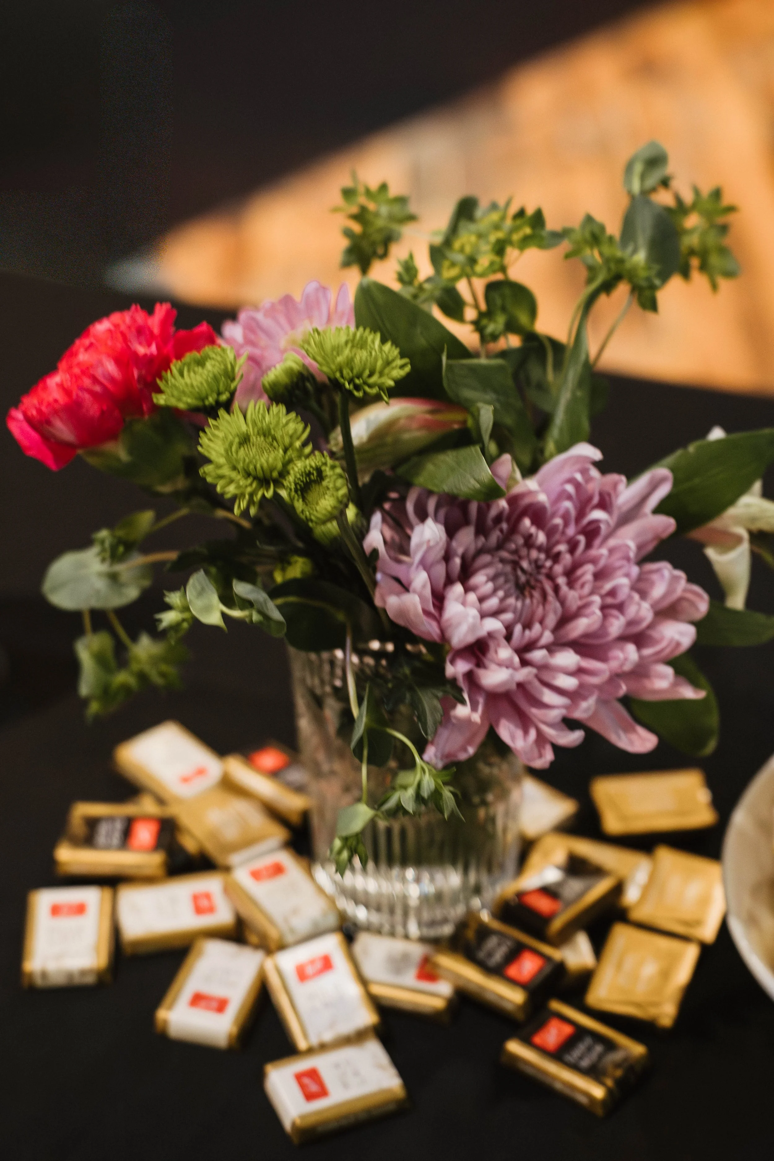 A bouquet of pink, red, and light purple flowers arranged in a glass vase, surrounded by scattered small, gold and white packages on a black table.