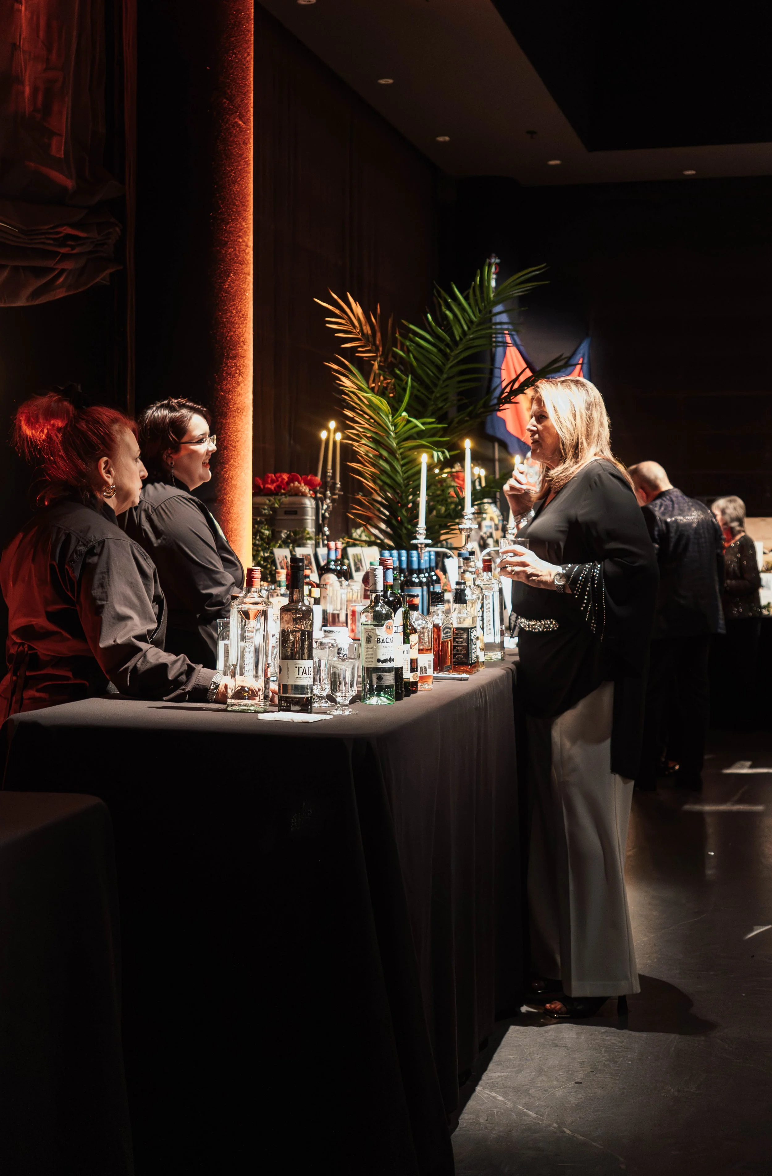 A woman in black and beige pants talking to two women behind a bar with bottles of alcohol, candles, and decorations. The scene is dimly lit with a dark background and some plants.