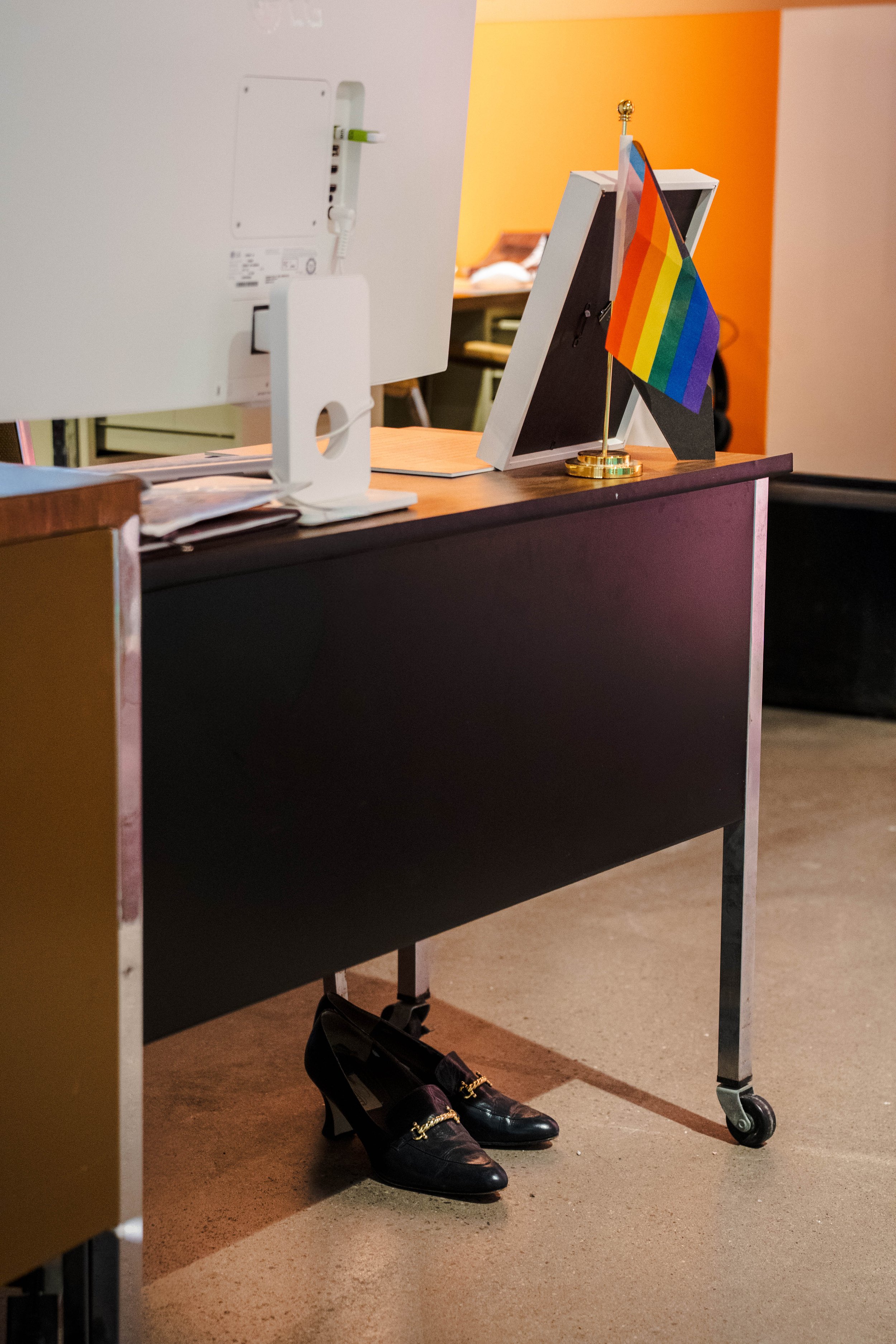 A desk with a computer monitor and a rainbow pride flag. Black high-heeled shoes are placed under the desk, and the background shows a room with a yellow wall.