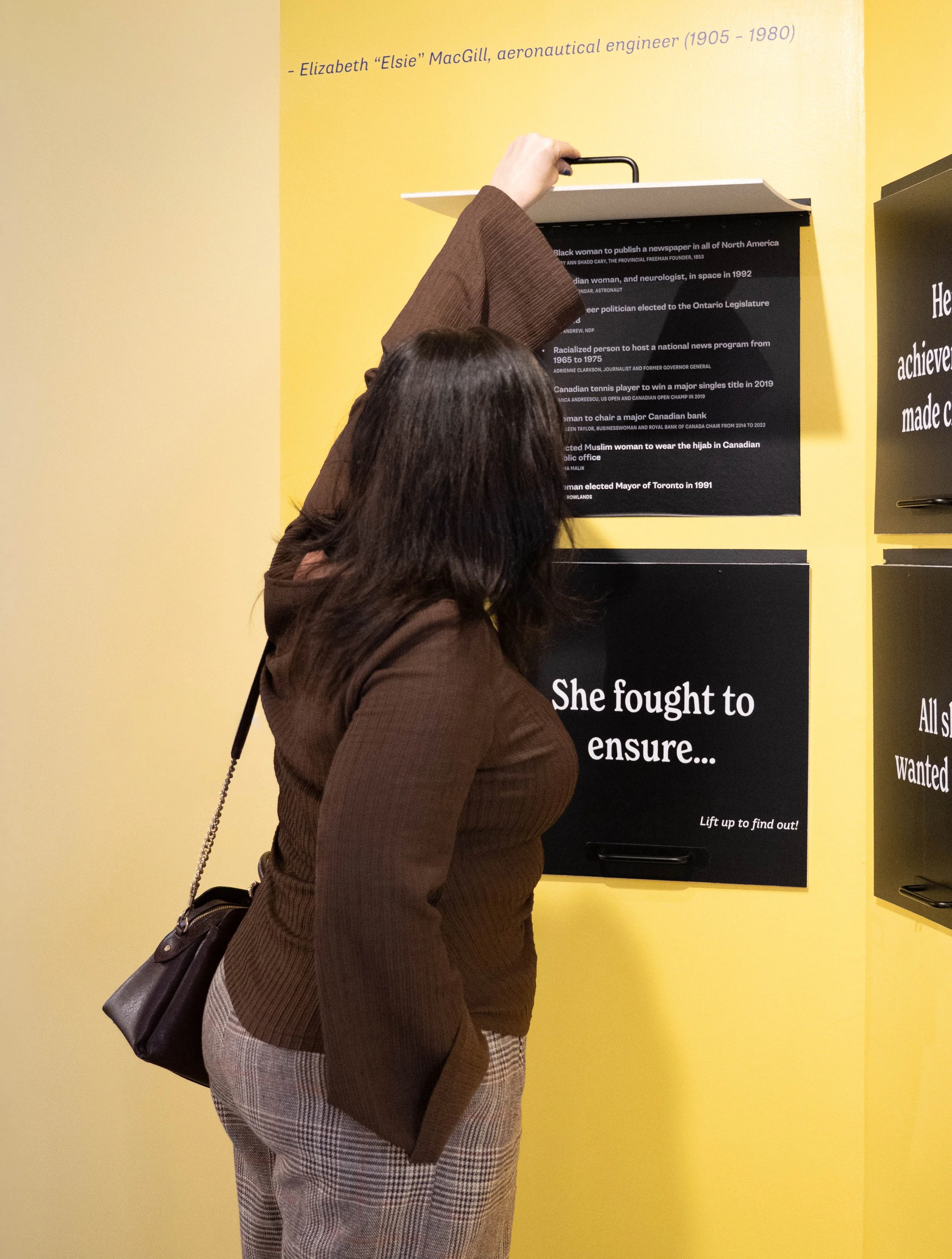 A woman in a brown sweater with plaid pants pulls a drawer open on a black display panel on a yellow wall in an exhibit or museum.