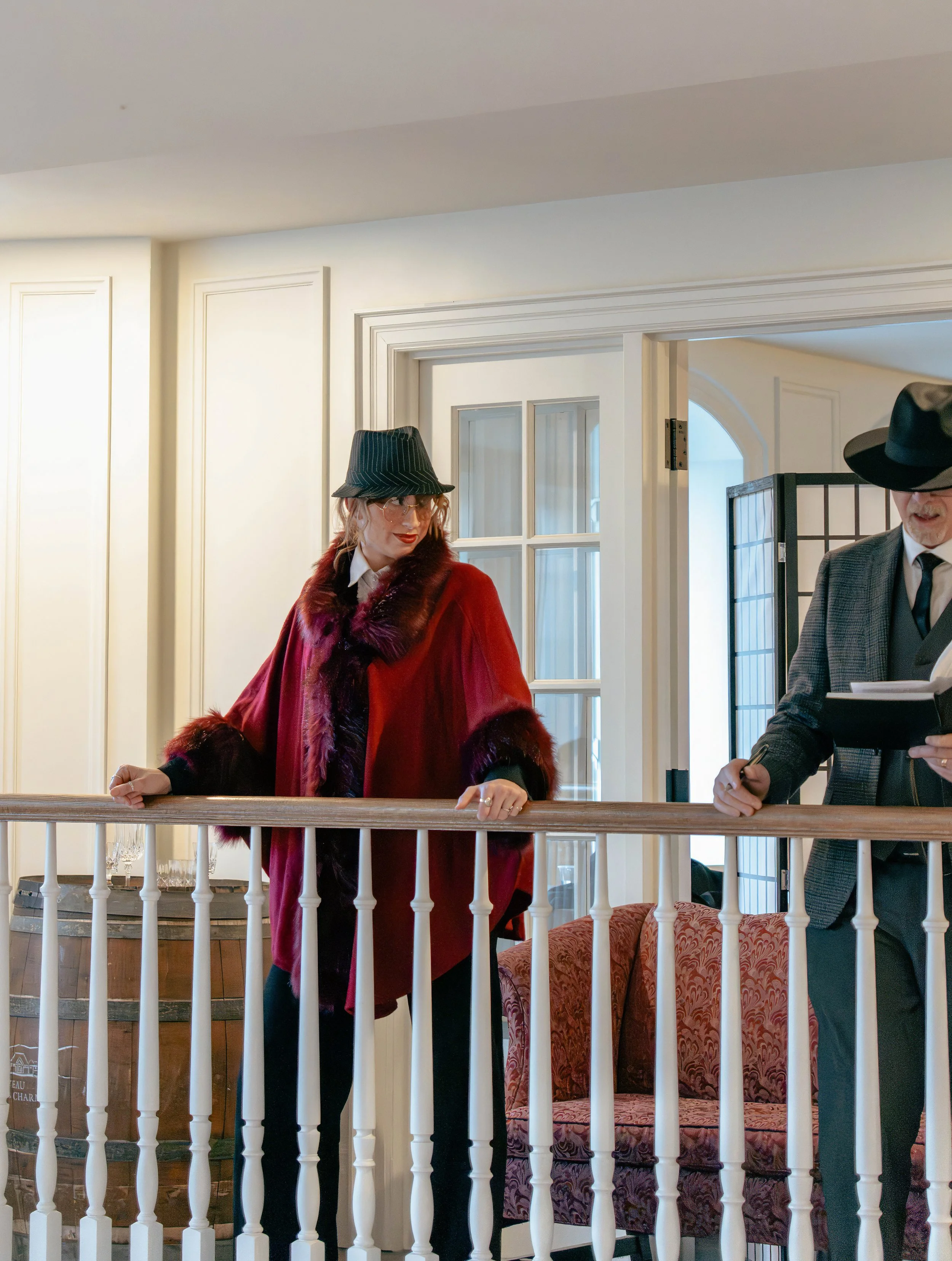 A woman in a red coat with fur trim and a black hat standing behind a white railing, with a man in a gray suit and large black hat nearby, in a bright room with white walls and a window.