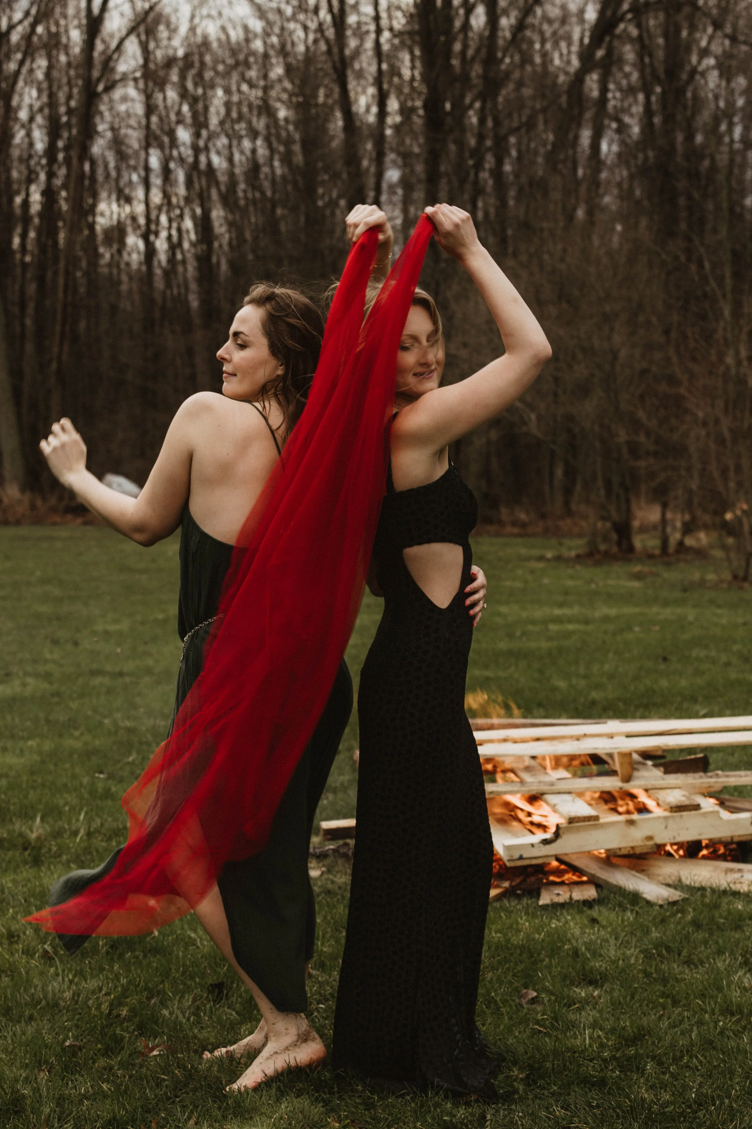 Two women dancing outdoors near a bonfire, wearing black dresses, one holding a red fabric, with trees in the background.