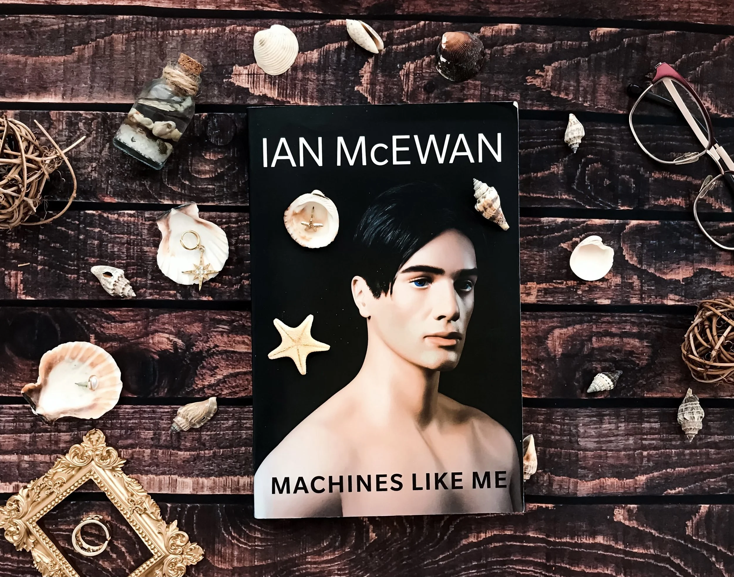 Book titled "Machines Like Me" by Ian McEwan placed on a dark wood surface surrounded by seashells, small decorative ball, and a pair of glasses.
