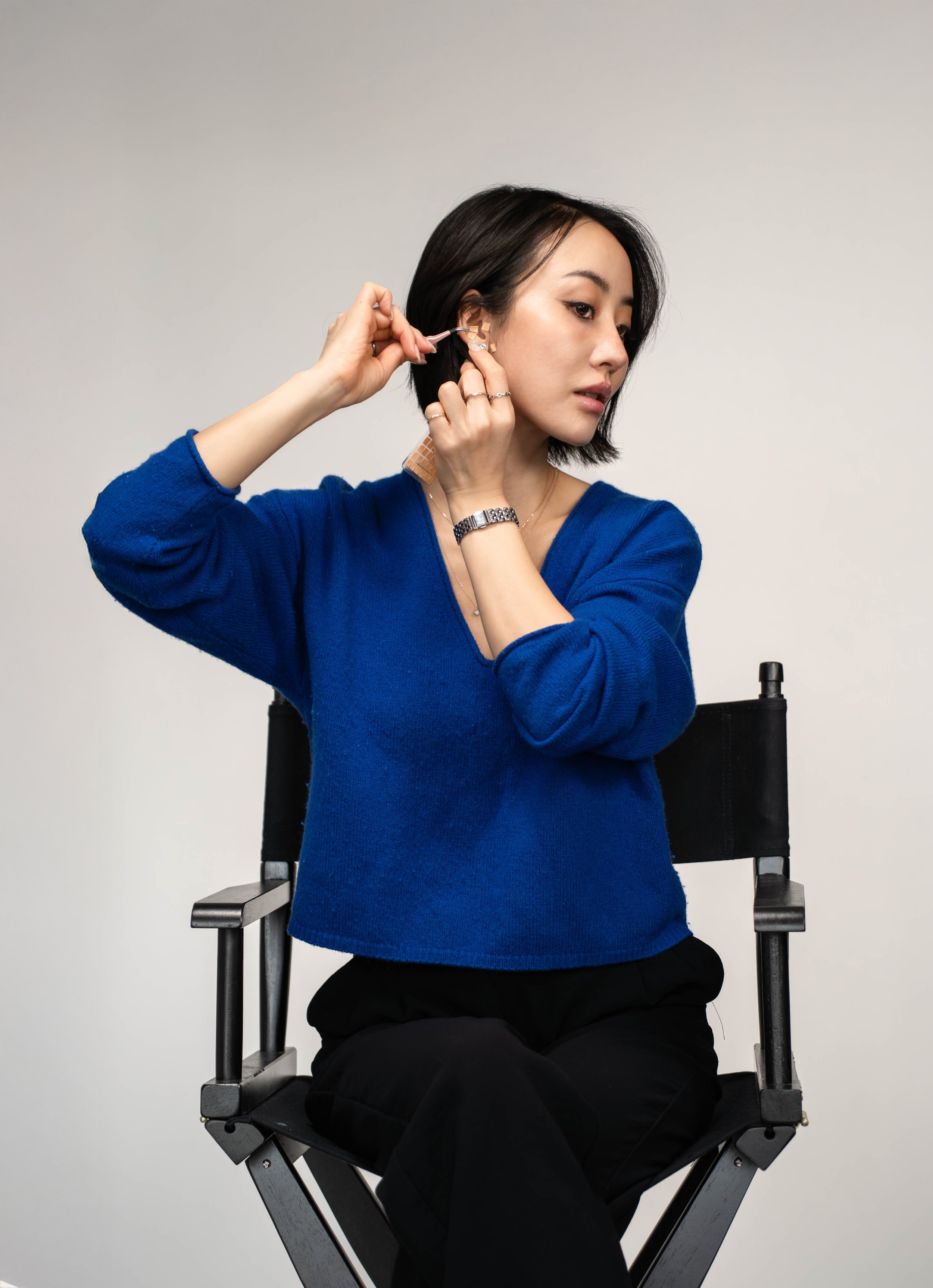 A woman with shoulder-length dark hair wearing a blue sweater and black pants is sitting on a director's chair, putting an earring in her right ear.