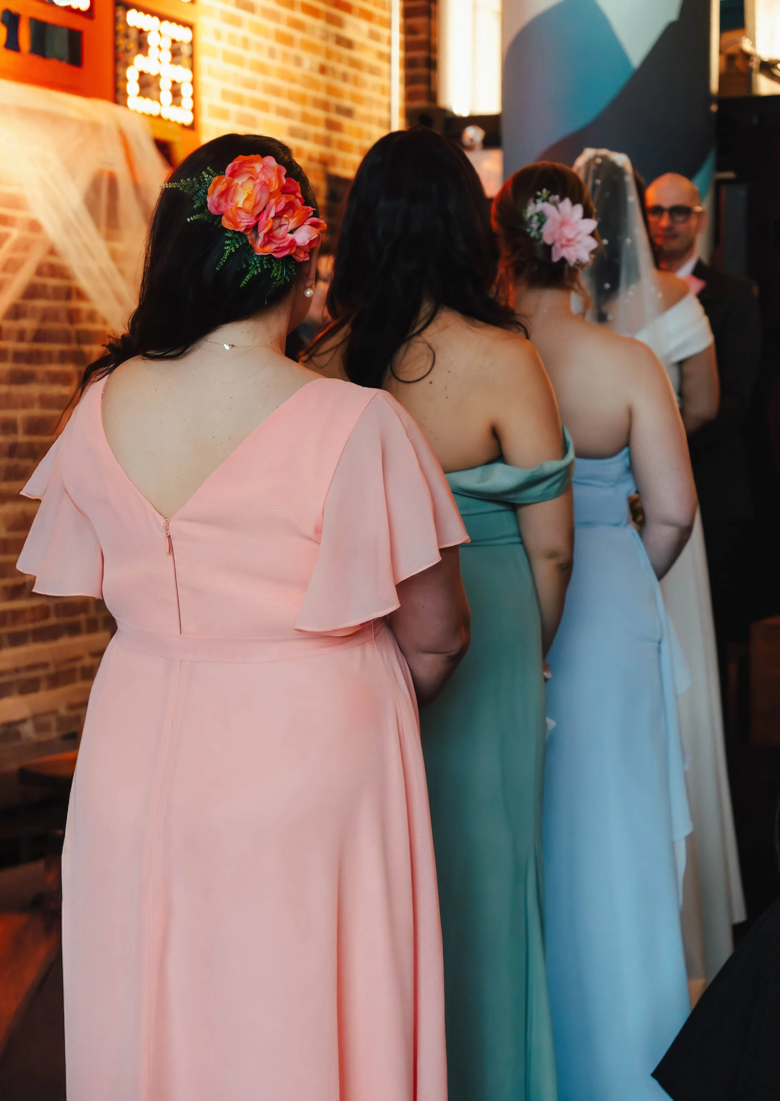 Line of women with floral hair accessories, wearing pastel-colored dresses, at a wedding ceremony.