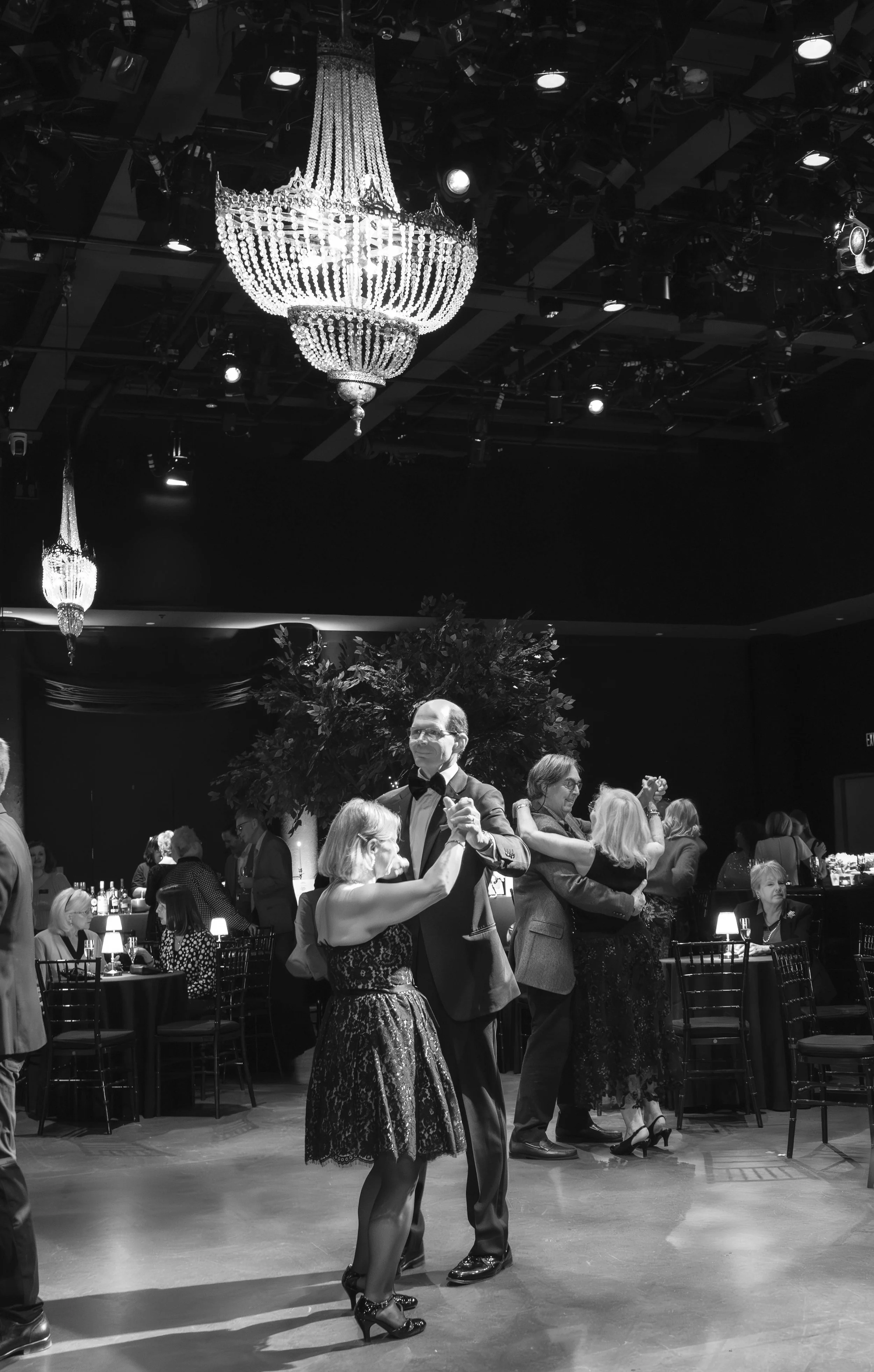 People dancing at a formal event under chandeliers in a decorated ballroom.